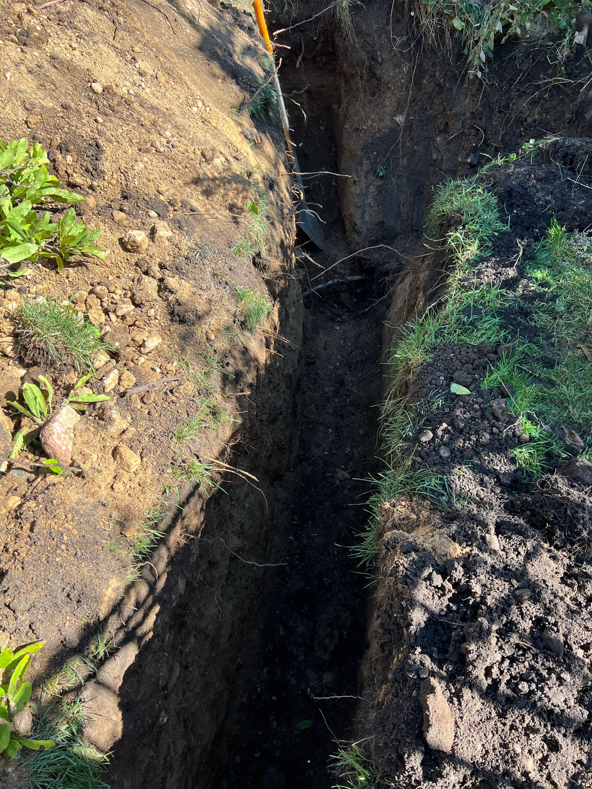 A narrow, deep trench dug into the earth, with exposed dark soil walls and patches of green plants along the edges.