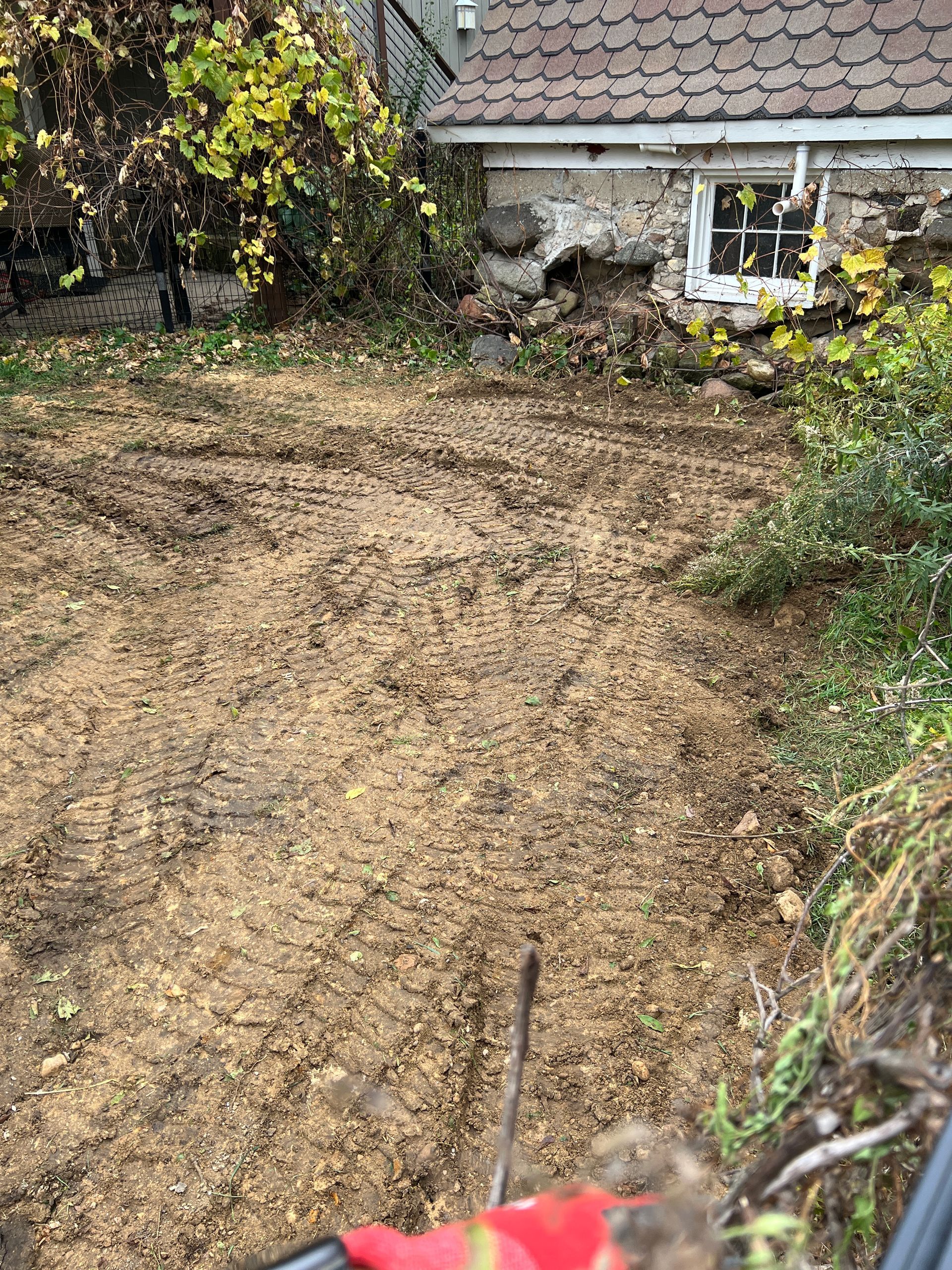 An area of recently tilled soil with tire tracks, set in front of a rustic stone building with a shingled roof.