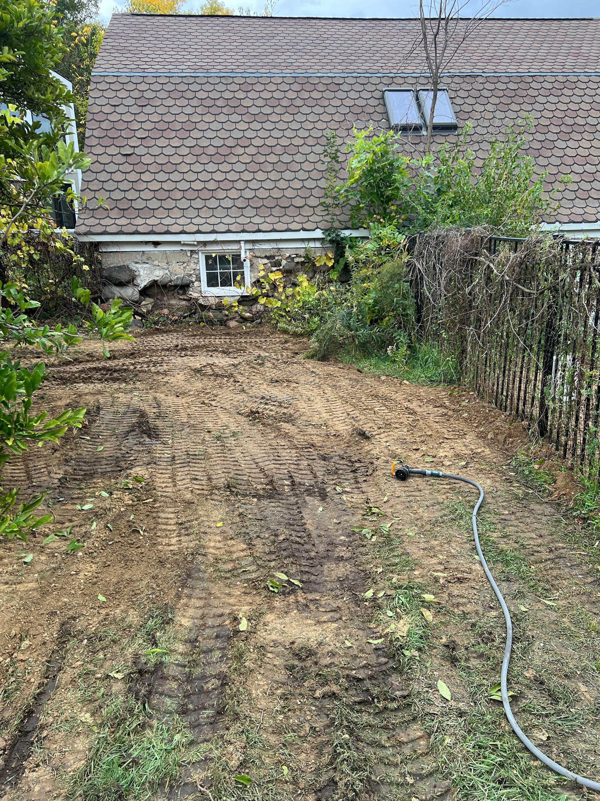 A patch of tilled, dirt-covered yard in front of a stone-foundation house with a large, shingled roof.