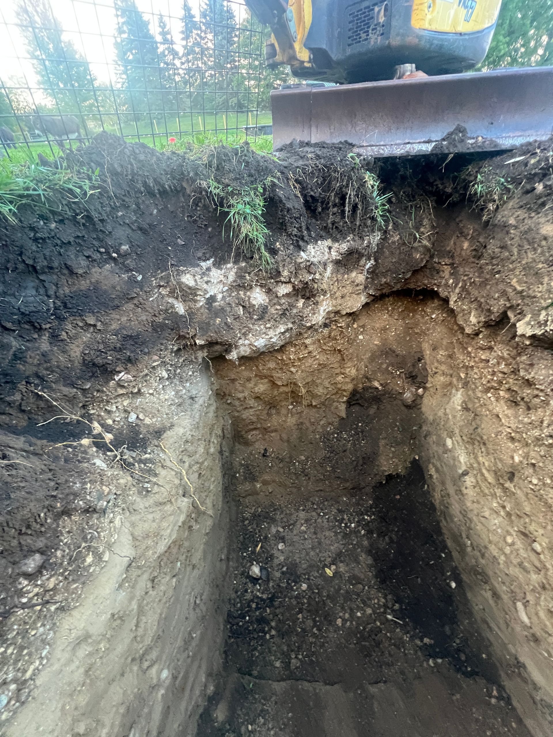 An excavator's blade sits atop a deep, rectangular hole dug into the earth, showing layers of dark and light soil.