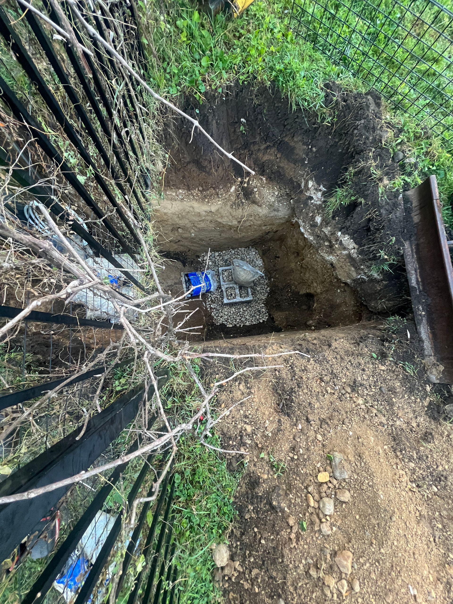 A square excavation pit in the ground next to a metal fence, revealing a utility valve with blue components.