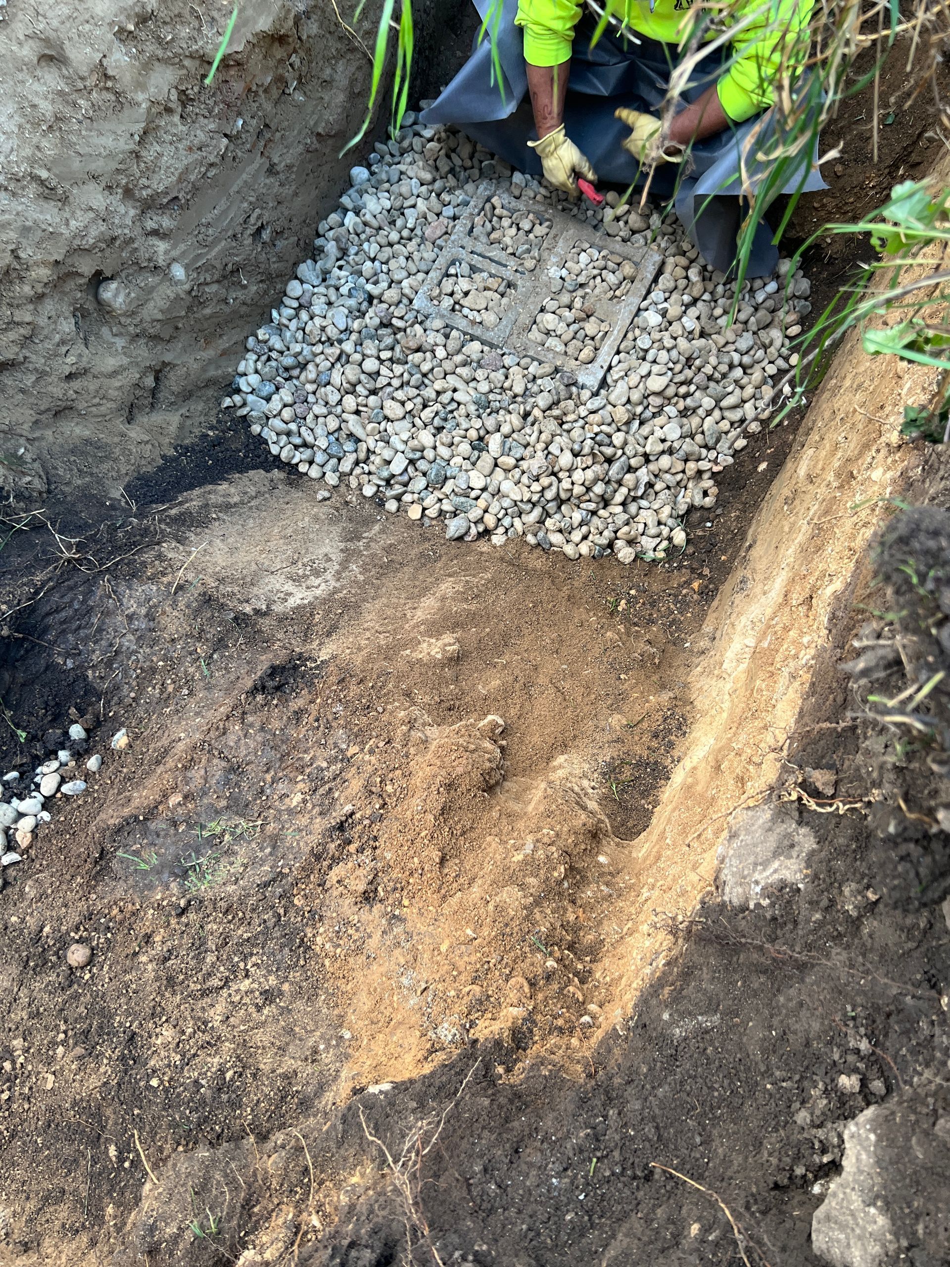 A person in high-visibility clothing uses a hand tool to spread gravel in a narrow dirt trench.