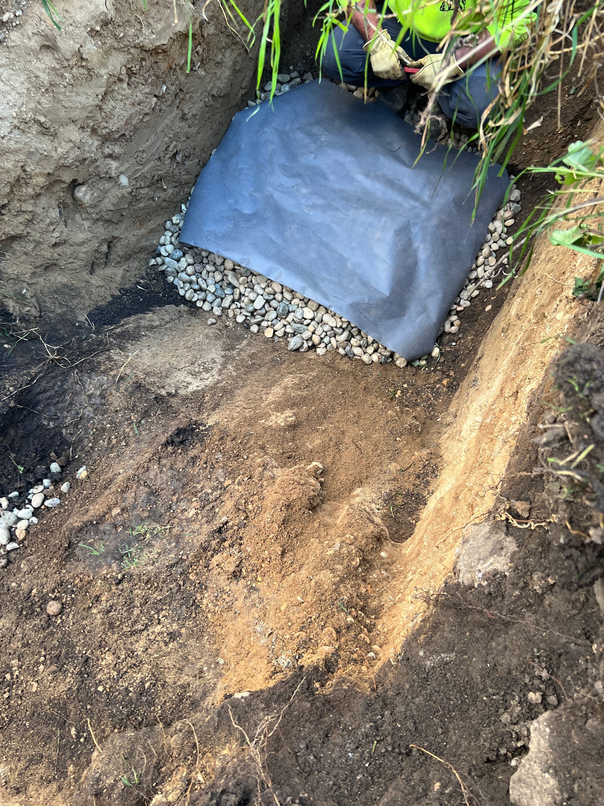 A view of a drainage trench under construction, showing gravel covered by black landscape fabric in a dirt excavation.