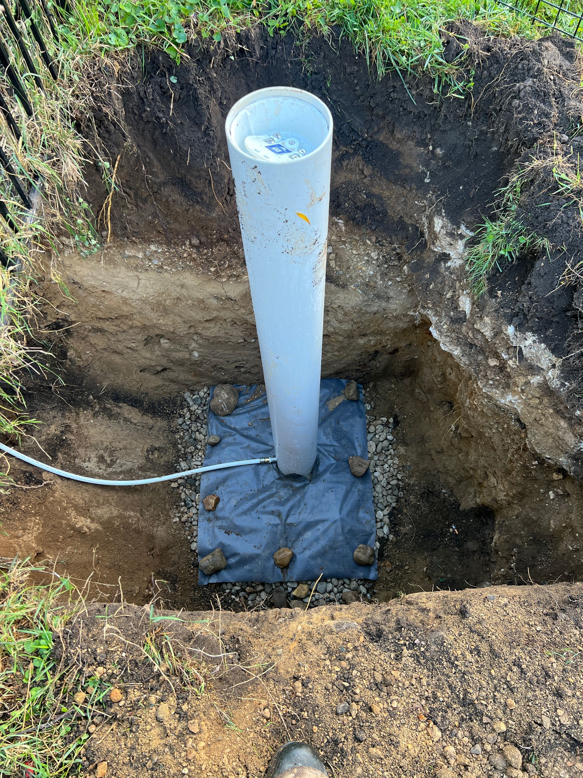 A white PVC pipe stands vertically in a hole in the ground, anchored on a gravel base covered by a black landscape fabric.