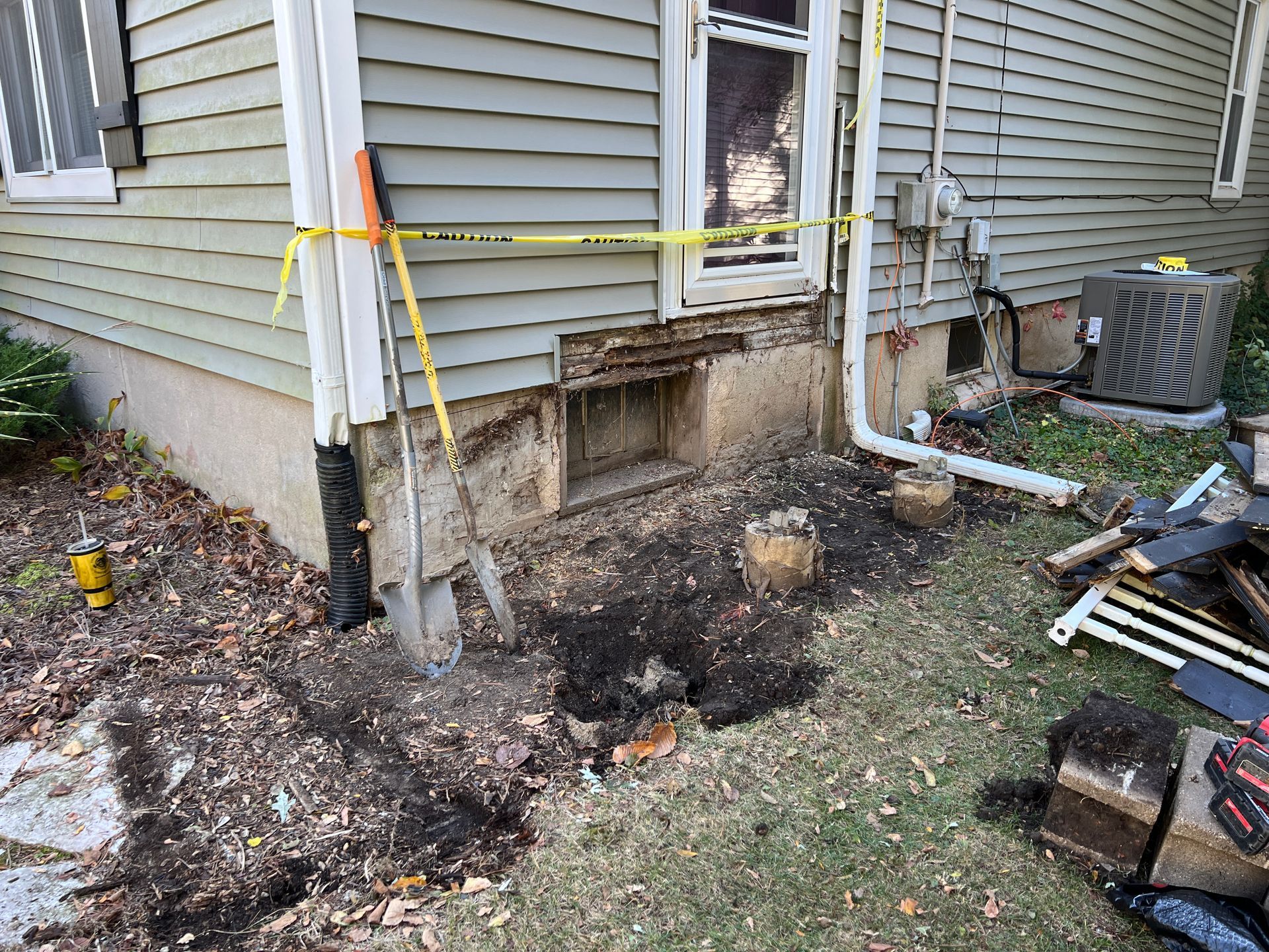 Exterior view of a house foundation with exposed concrete, excavation work, and caution tape marking the area.