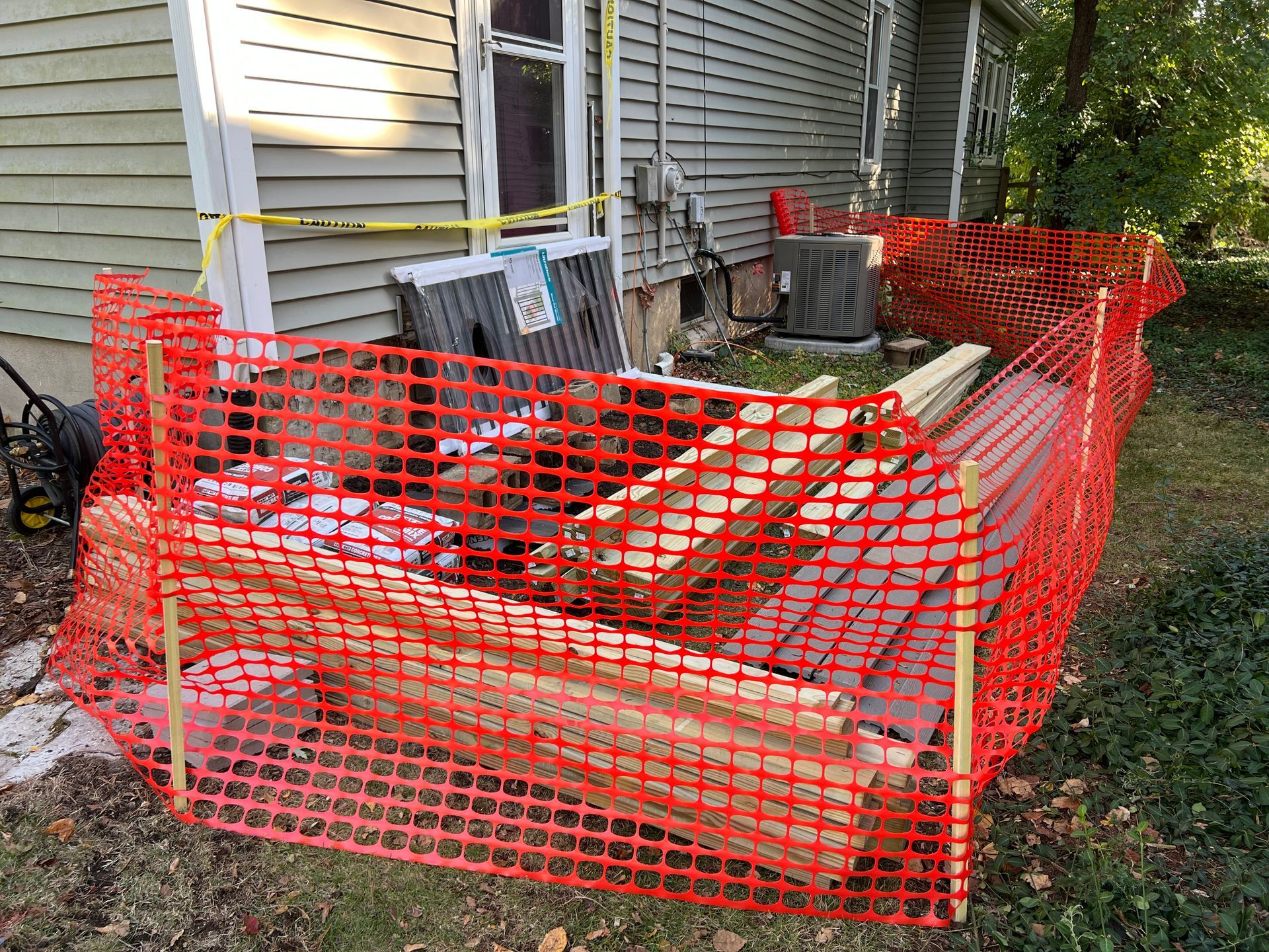Construction site materials, including wood boards, enclosed by orange safety fencing next to the side of a house.