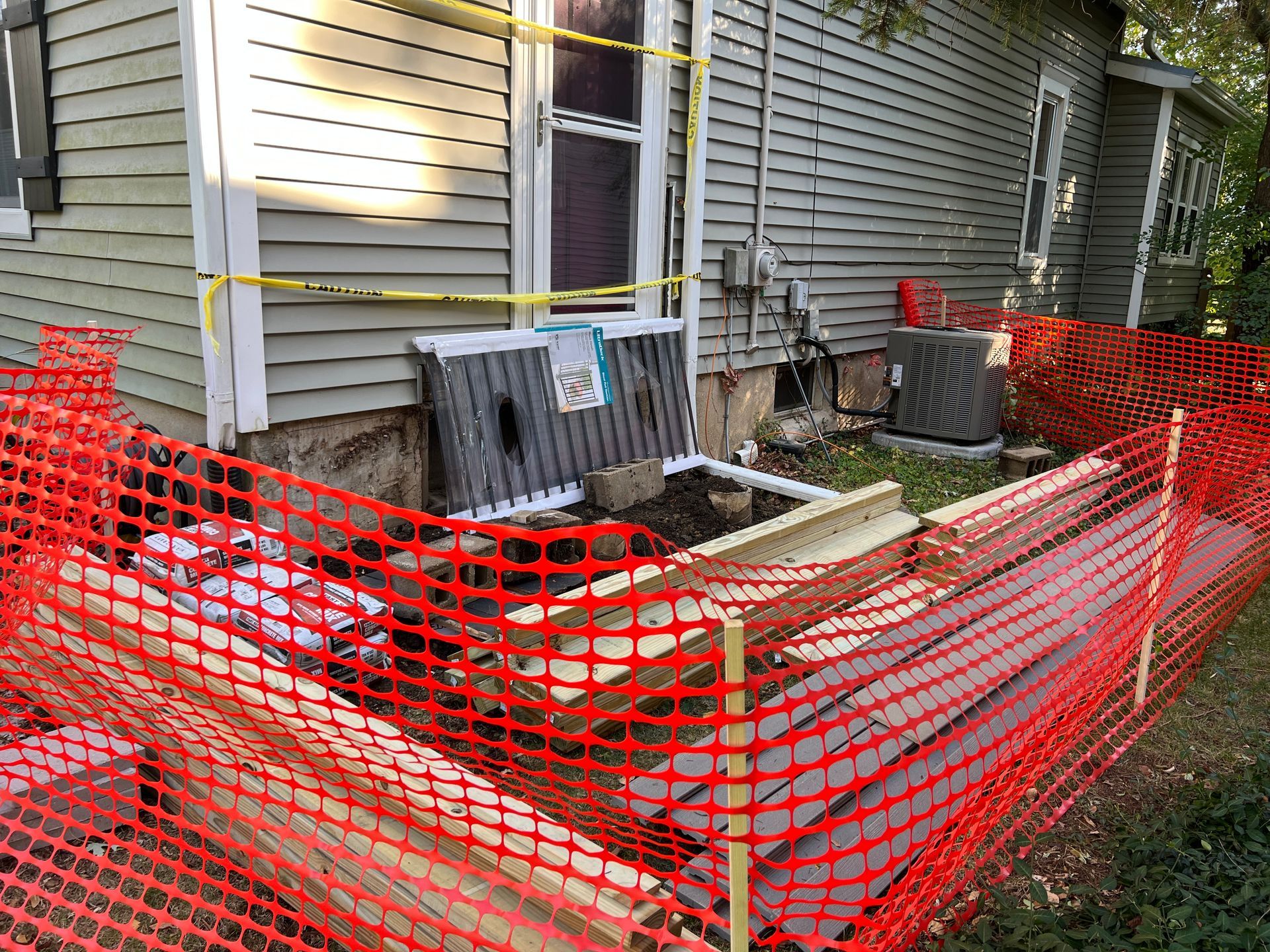 Construction site outside a house showing an open foundation pit surrounded by orange safety fencing and caution tape.