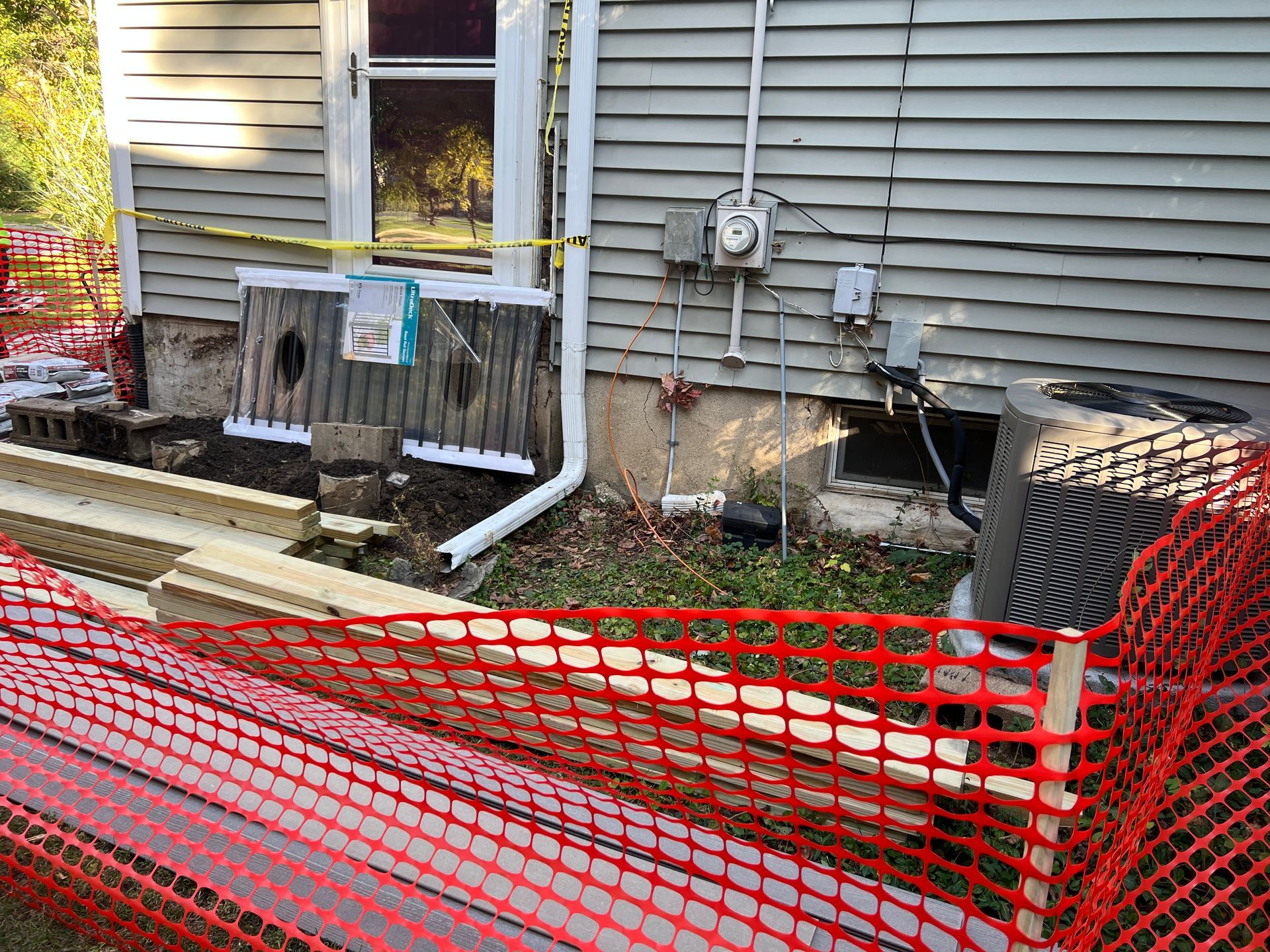 An outdoor construction site featuring exposed foundation work, wooden beams, and orange safety fencing against a house.