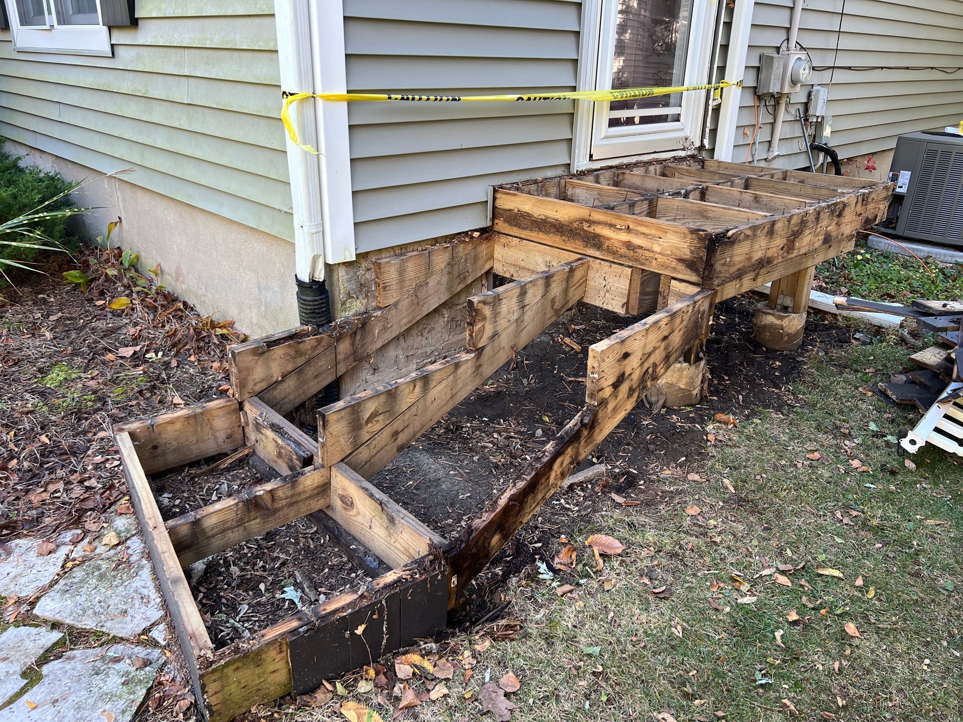 A side view of a house deck with the decking removed, exposing the wooden structural frame and yellow caution tape.