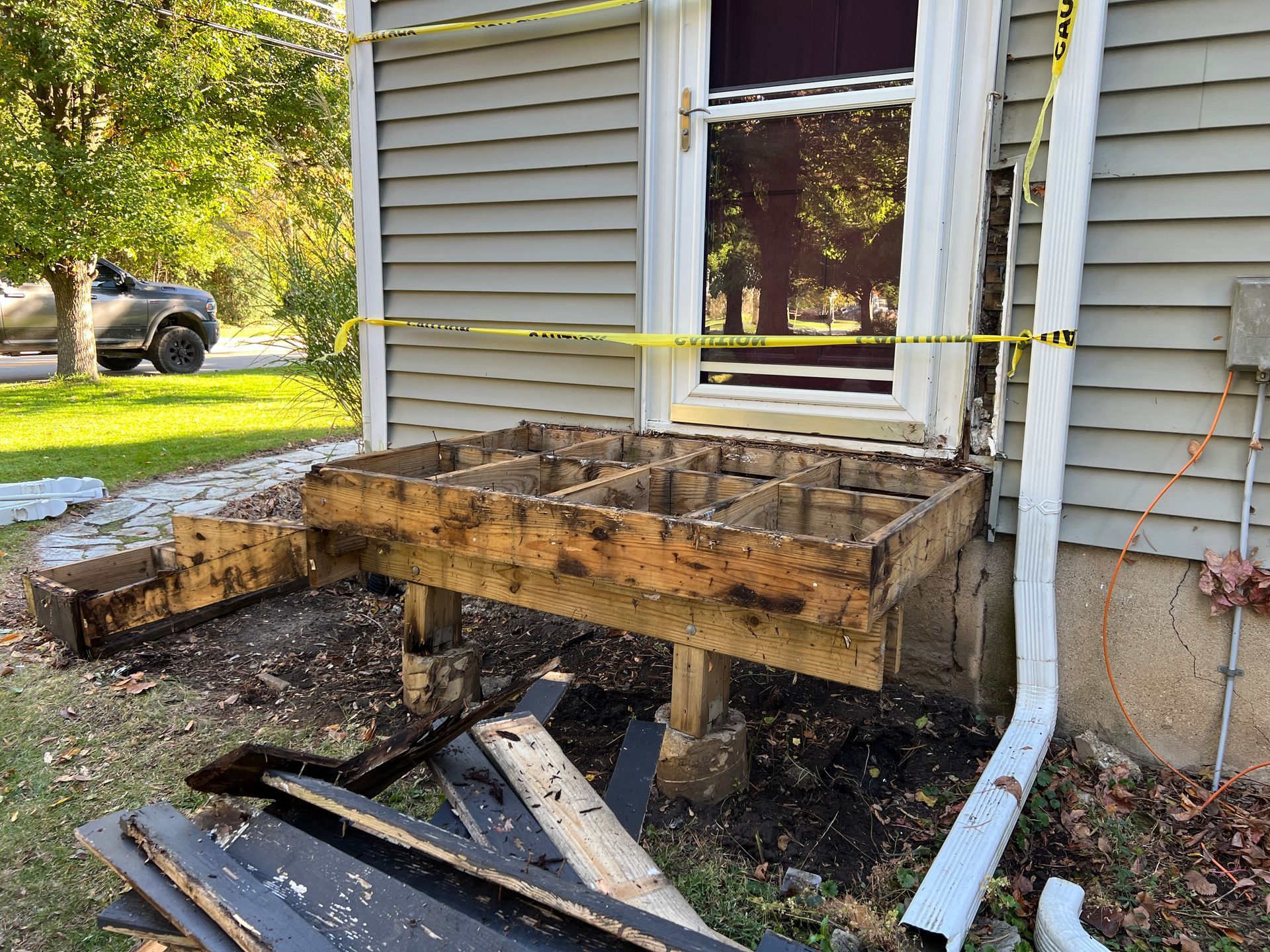 A damaged wooden deck frame stripped of its surface boards, cordoned off by yellow caution tape against a gray house.