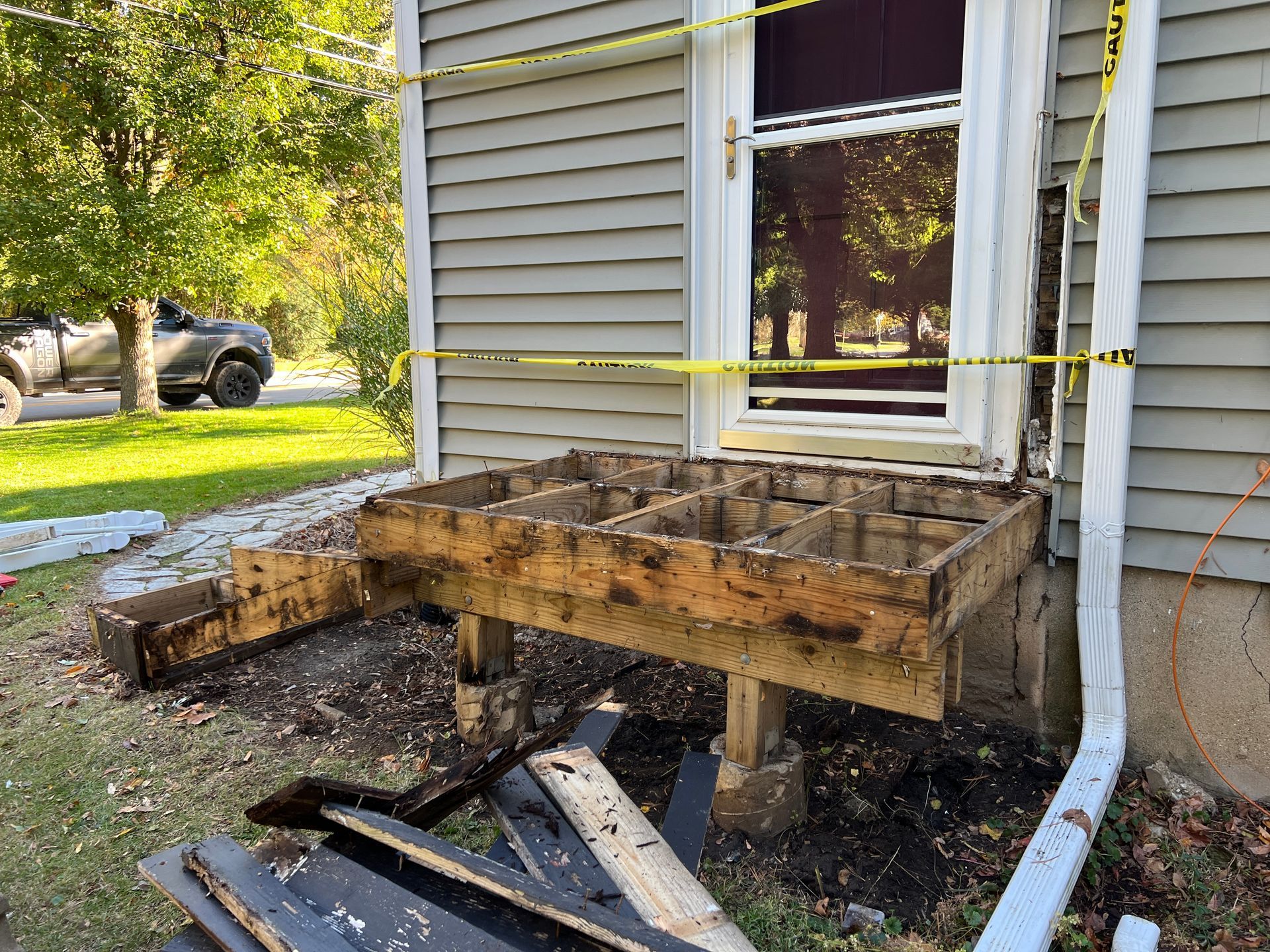 A porch with its decking removed, exposing the wooden frame and support posts, wrapped in yellow caution tape.