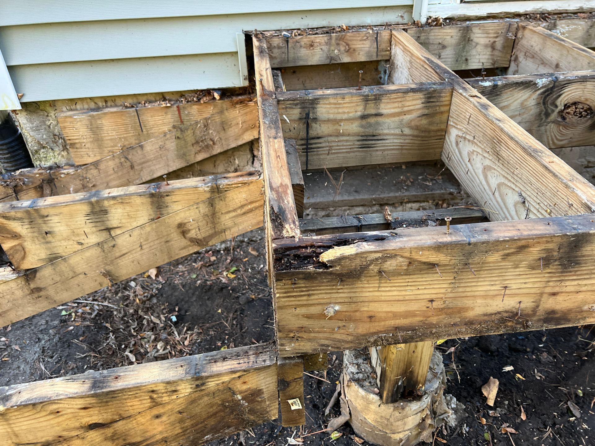 A weathered wooden deck frame with rot and damage, exposed to dirt, next to a house exterior.