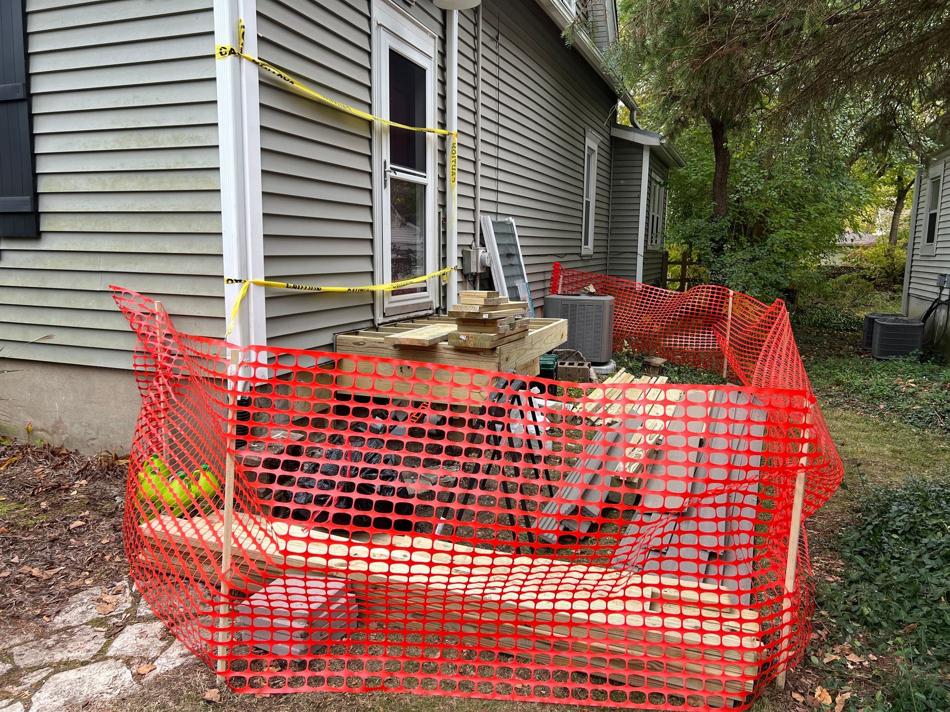A house exterior with construction work, featuring a wooden platform behind orange safety fencing and yellow caution tape.