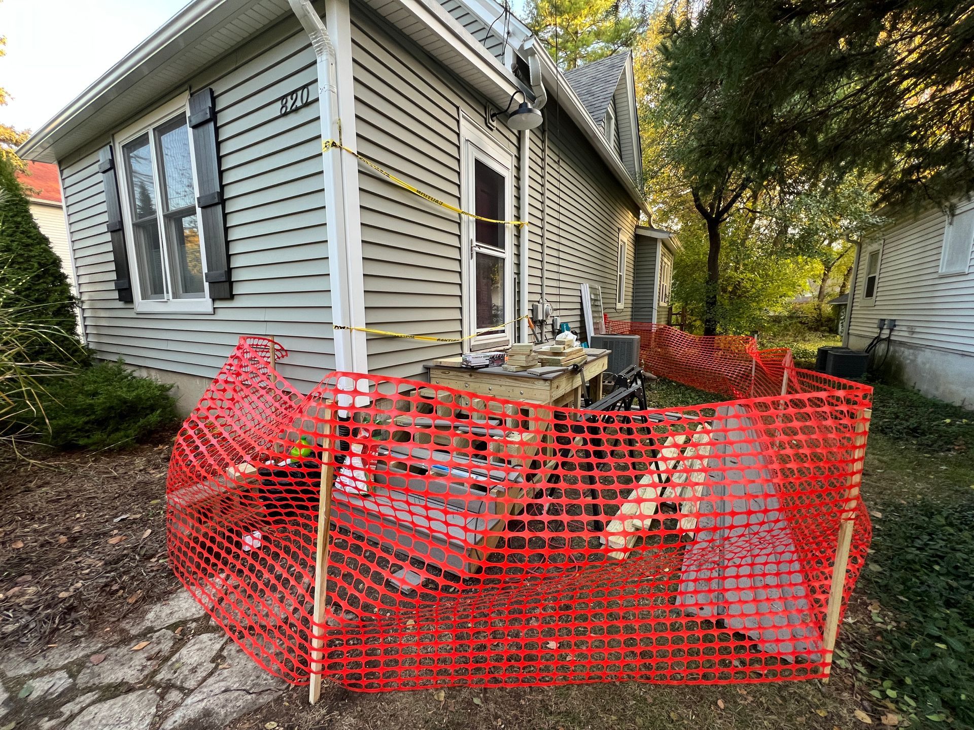 A grey house exterior with a wooden deck under construction, surrounded by a bright orange safety fence and caution tape.
