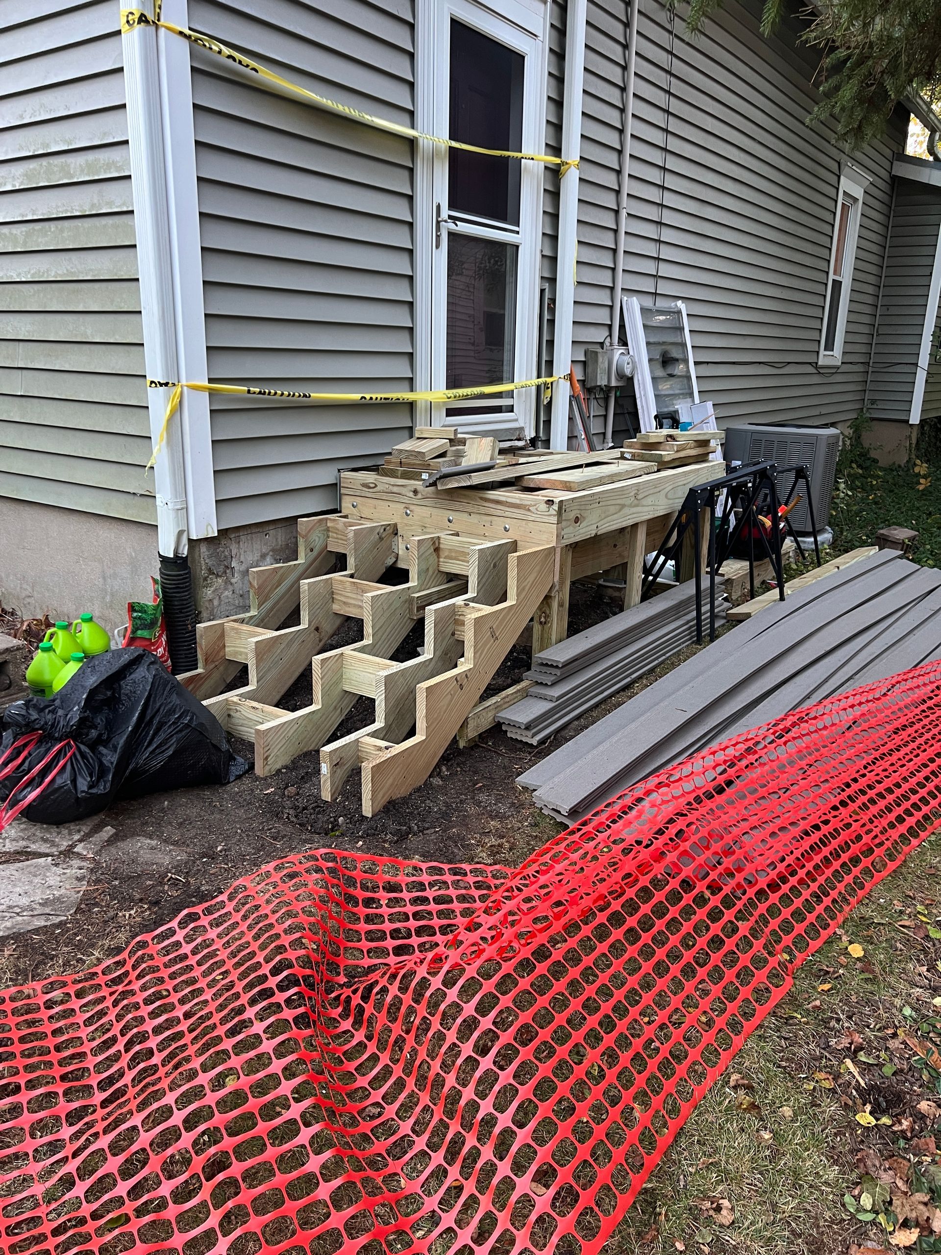 New wooden stairs under construction outside a gray house, with materials and safety fencing nearby.