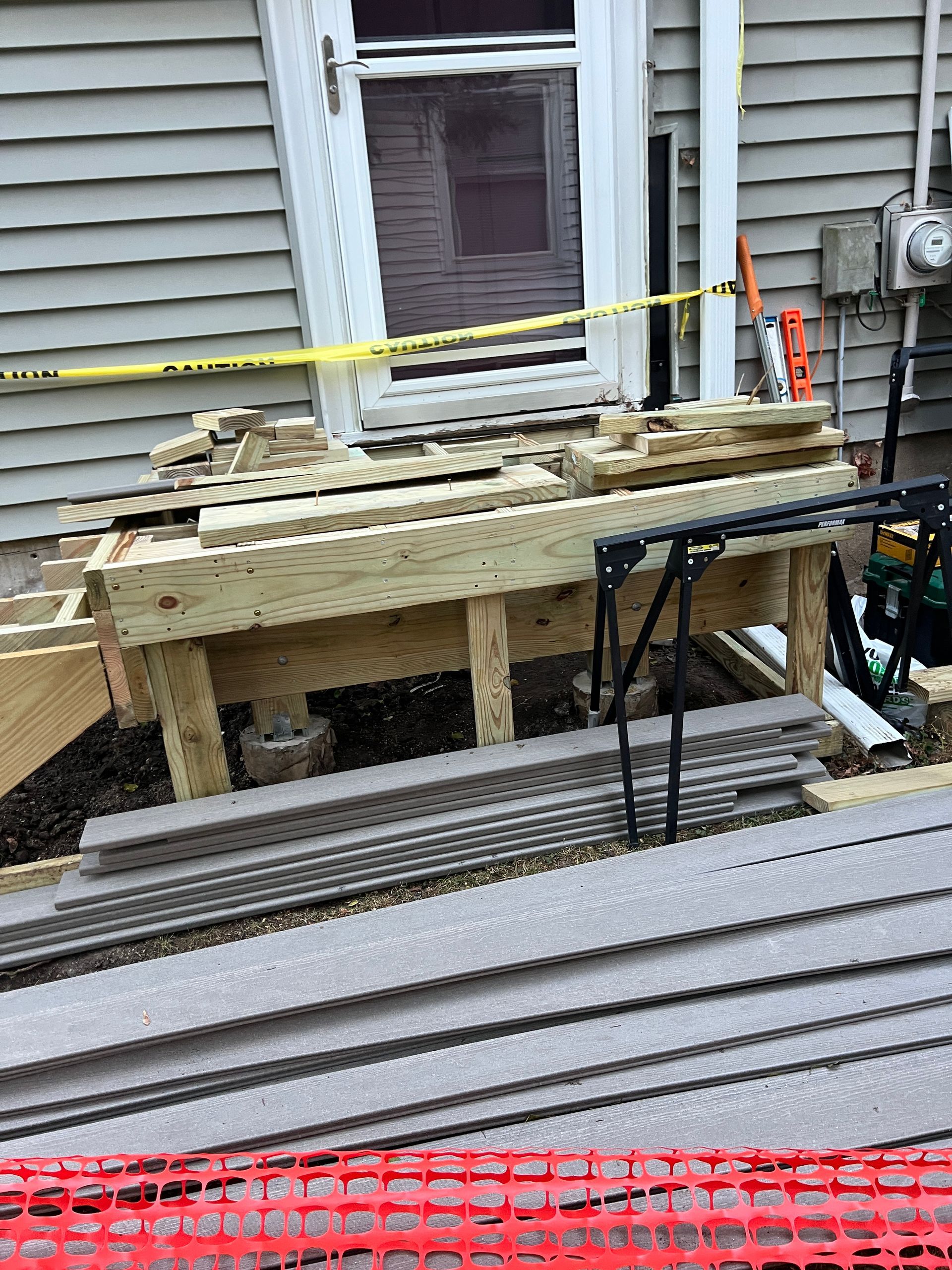 Construction of a wooden deck frame in progress outside a house, with gray decking materials stacked in the foreground.