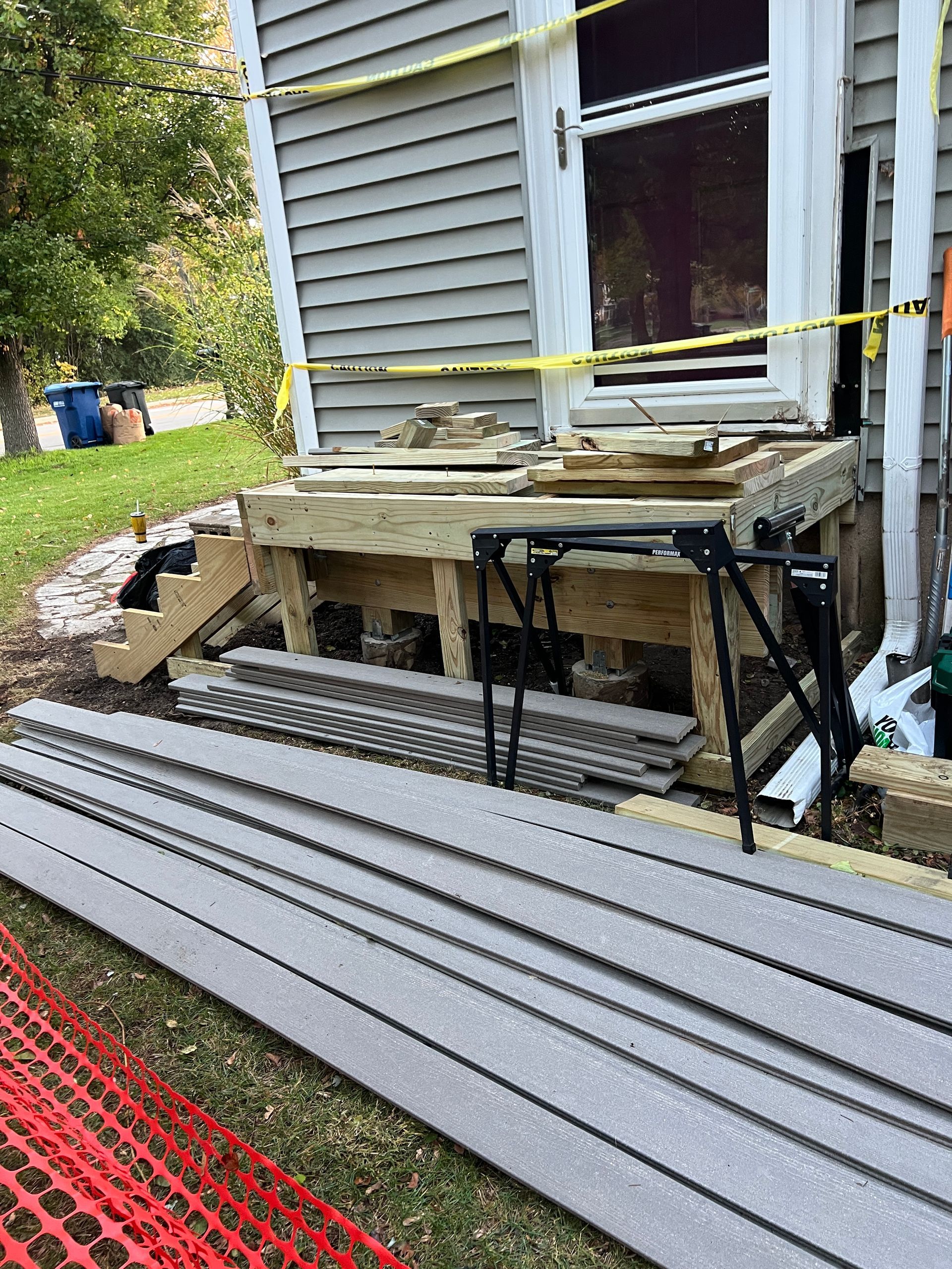 Construction work on a gray house exterior, featuring a wood deck frame and stacks of siding boards on the lawn.