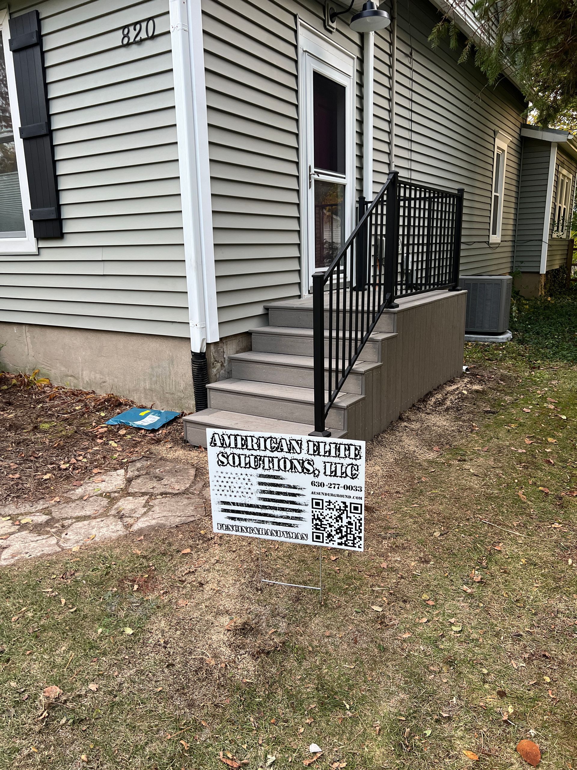 A lawn sign with an American flag graphic stands in front of a house with grey siding and a black front porch railing.
