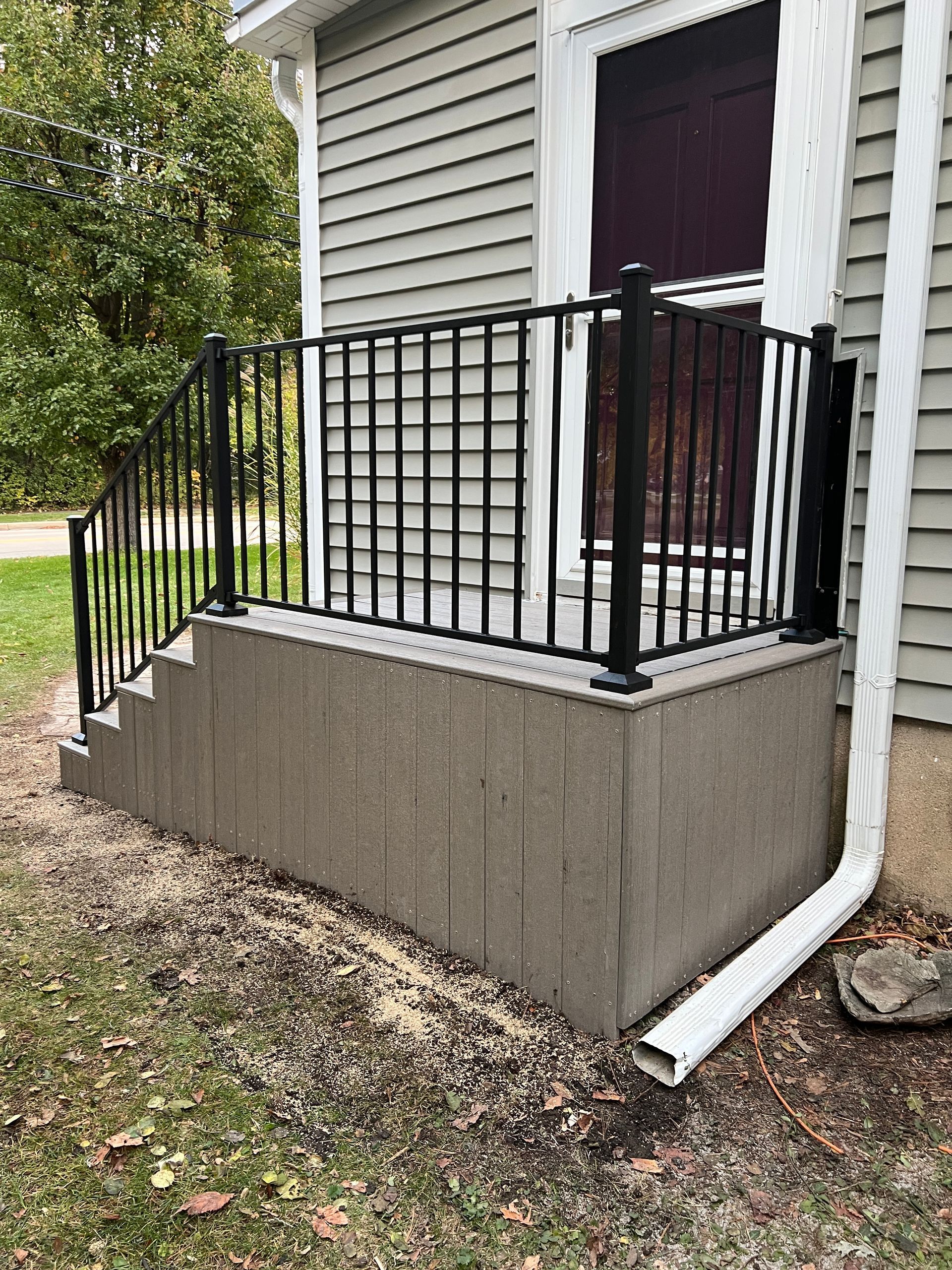 A concrete porch with black metal railings leading to a house door, with a white downspout on the right.