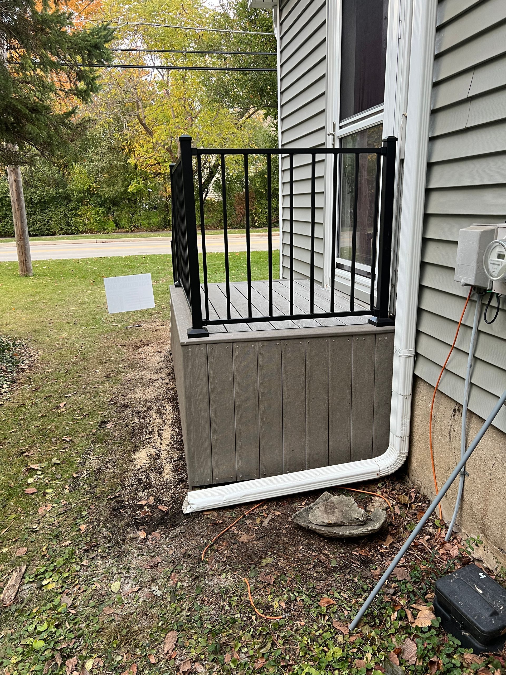 A small wooden deck with black metal railings next to a grey house, featuring a white gutter running along the base.