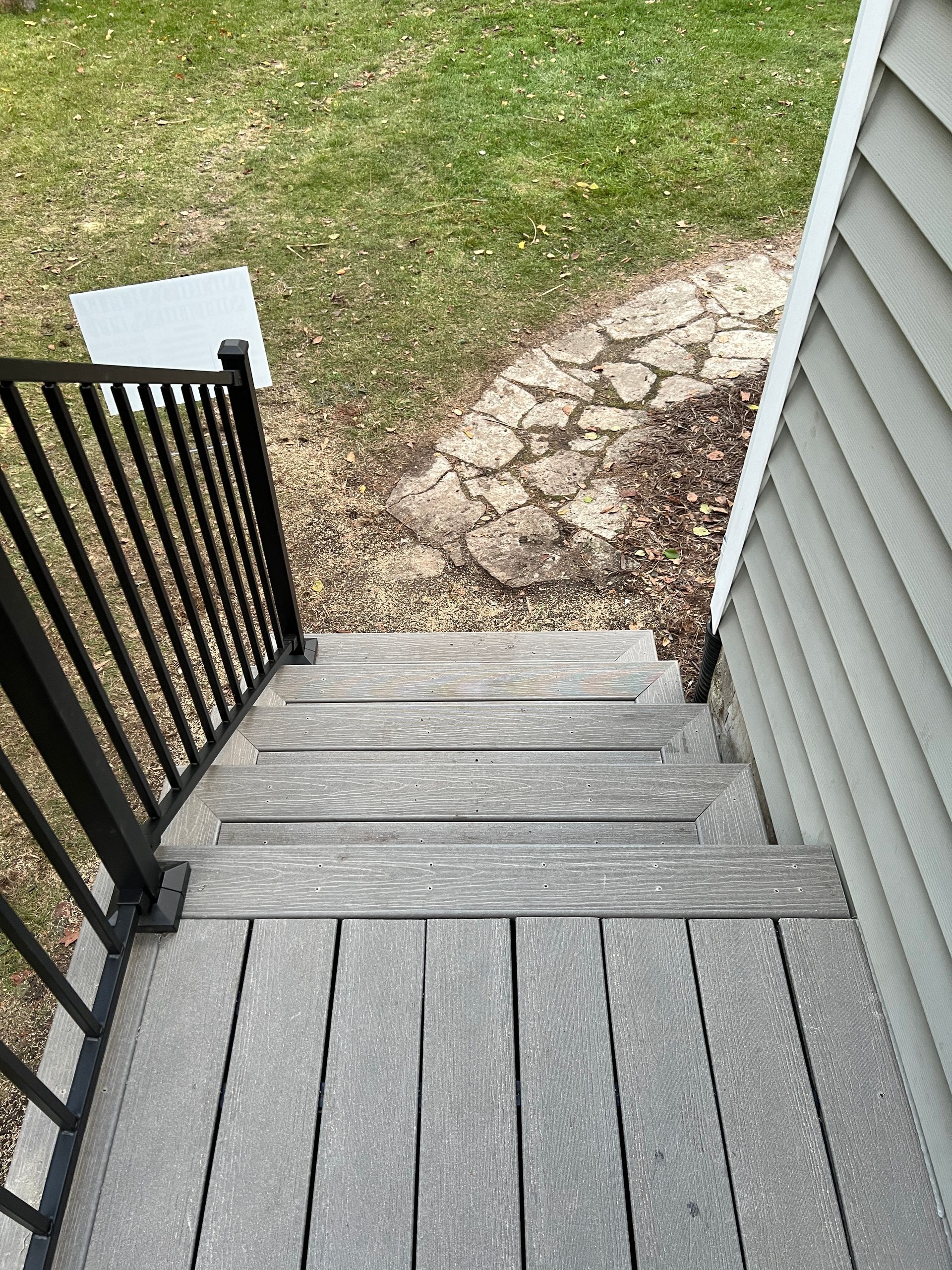 A high-angle view of gray deck stairs descending to a short stone path in a grassy yard, with a black metal railing.
