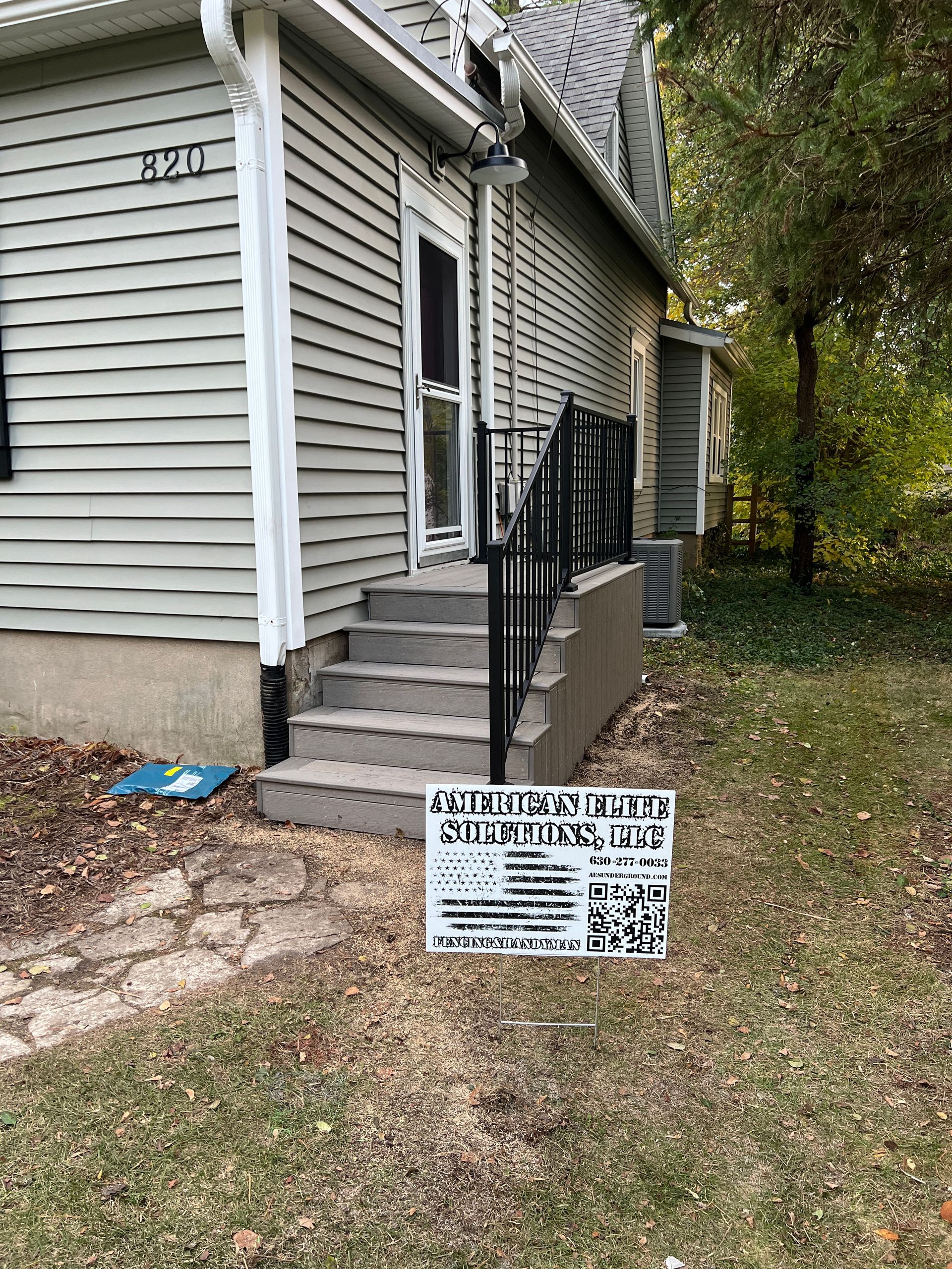 A light grey house with steps leading to a side door, featuring a sign with text and graphics on the lawn.