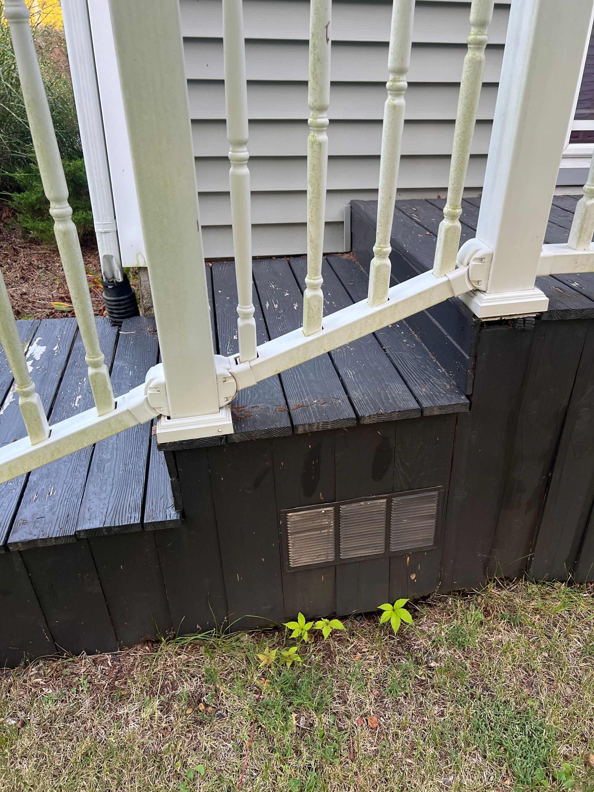 A white railing with spindles atop dark-painted wooden porch steps, featuring a small ventilation grate in the base.