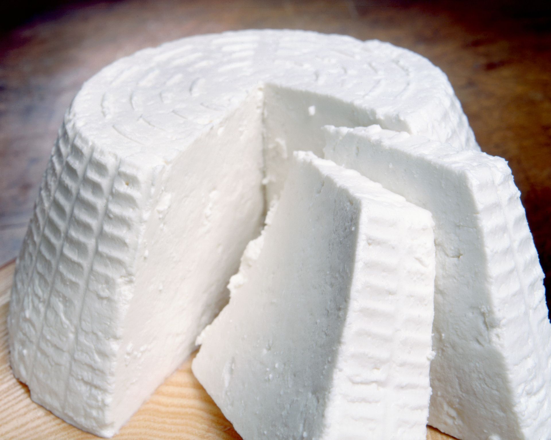 Ricotta bianca rotonda con uno spicchio tagliato e fette su un tagliere di legno.