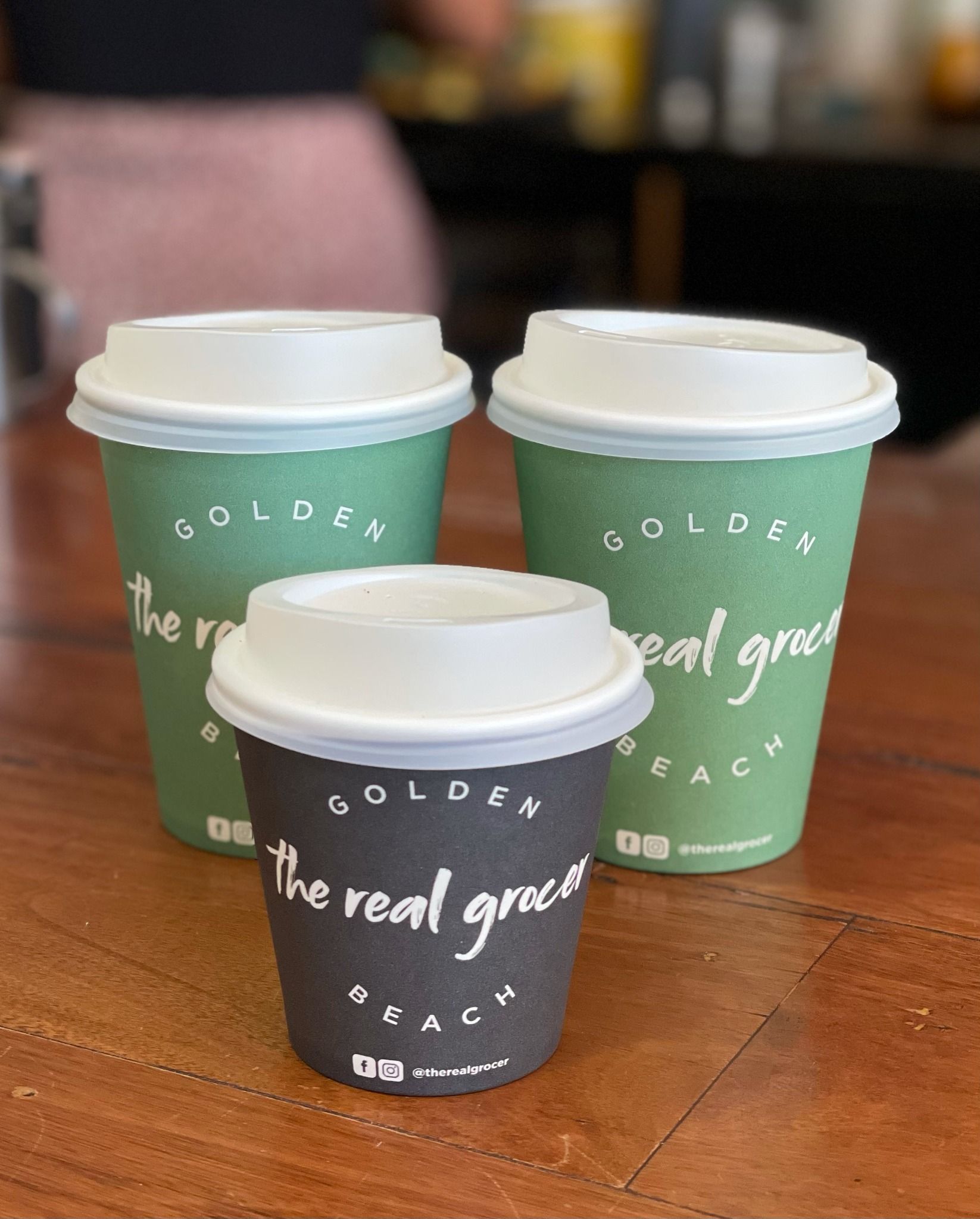 Three Paper Coffee Cups With White Lids, in Green and Black, on a Wooden Table — The Real Grocer In Golden Beach, QLD