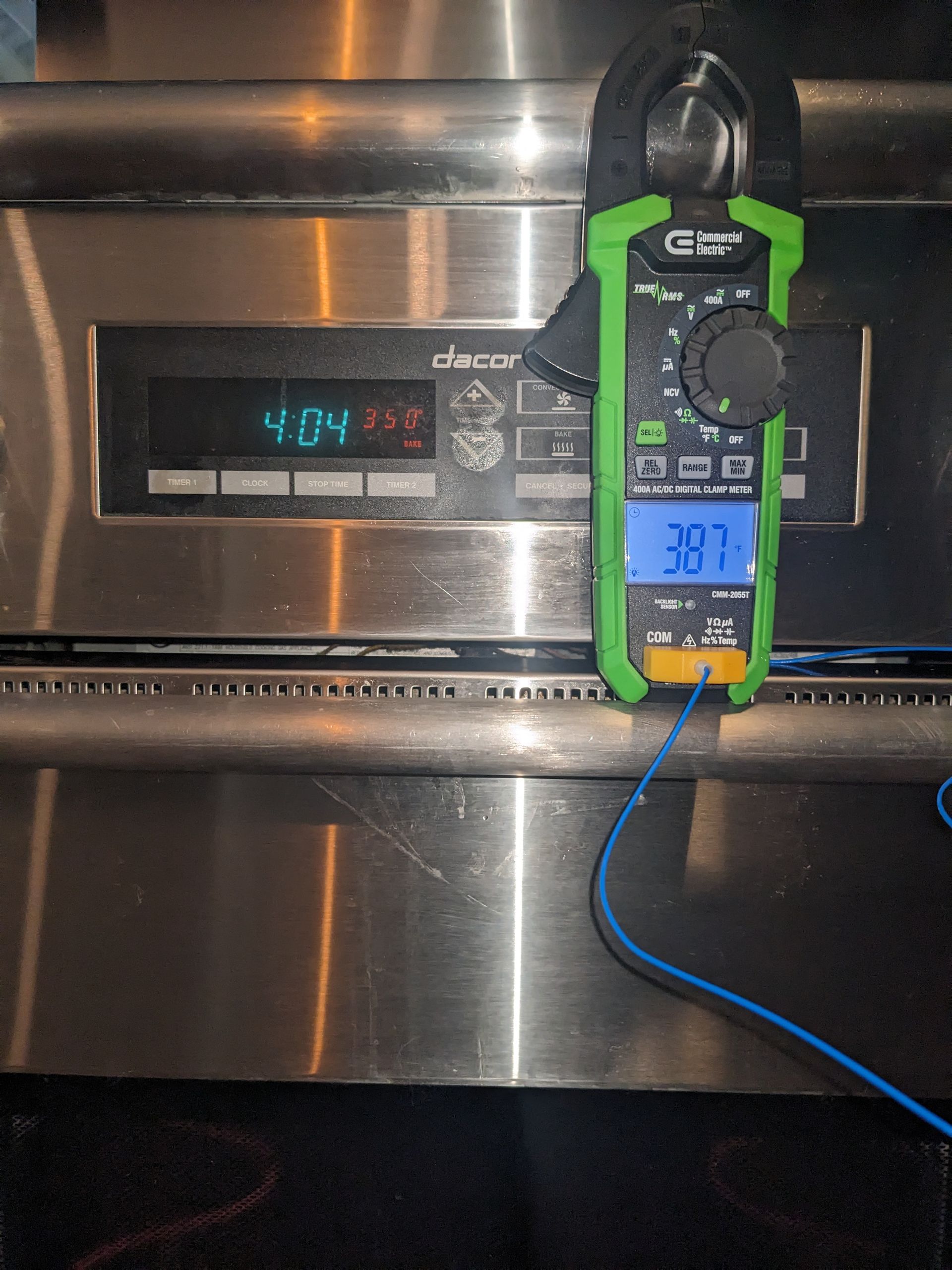 Multimeter clipped to a stainless-steel oven control panel, showing a temperature reading.