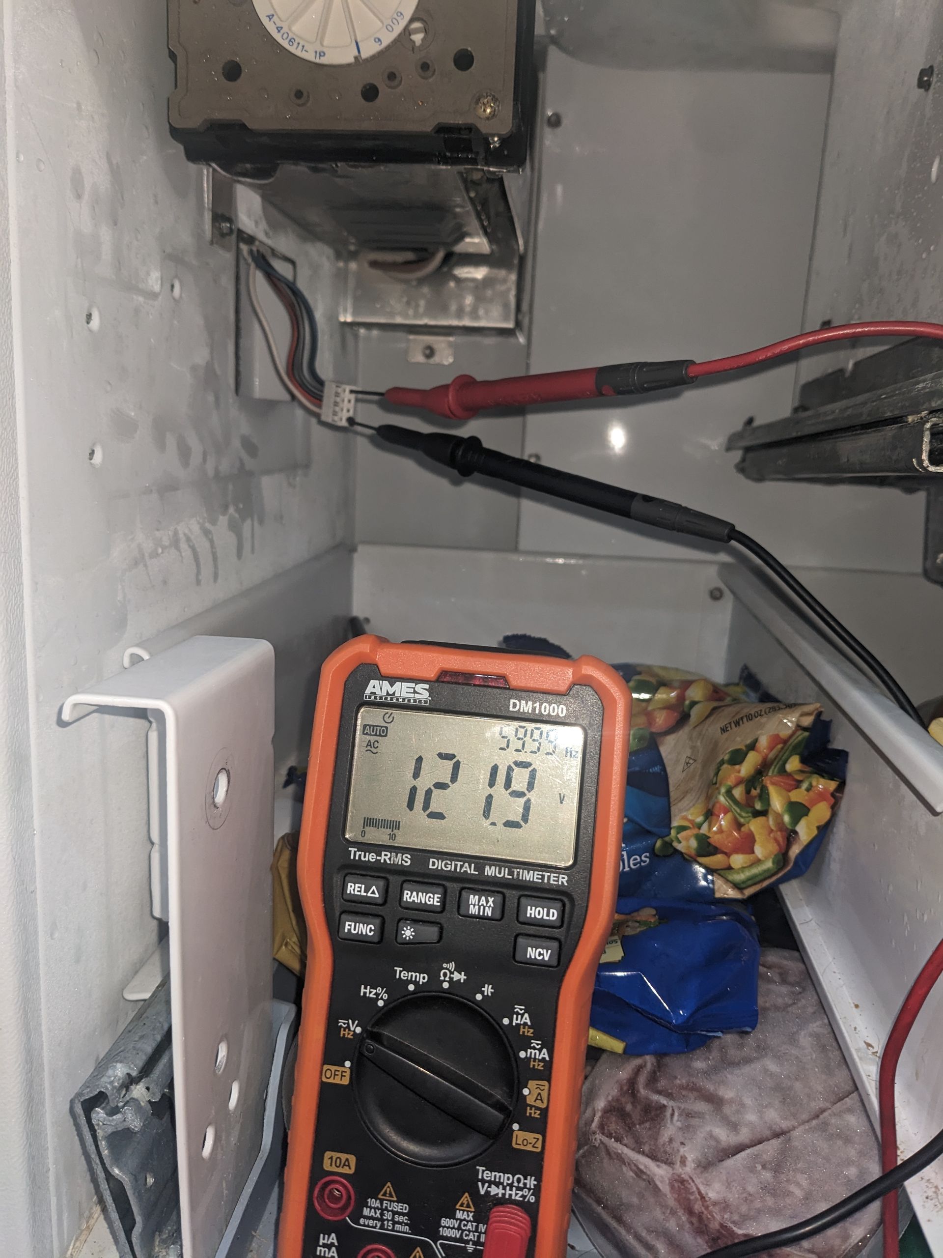 Multimeter inside an electrical panel reads 12.13 volts amid wiring and tools