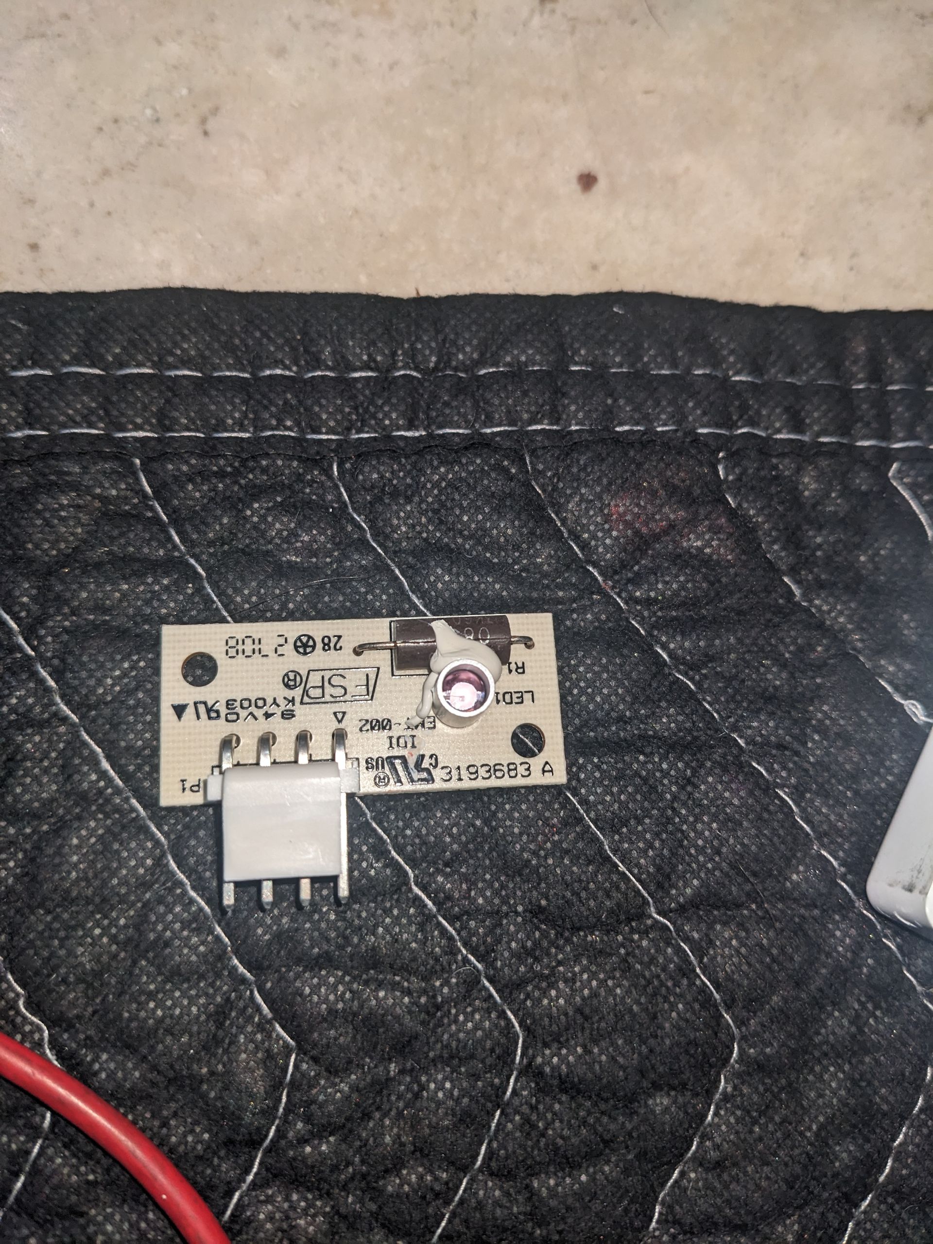 Small beige circuit board with a white connector on a black quilted surface, with a red wire nearby