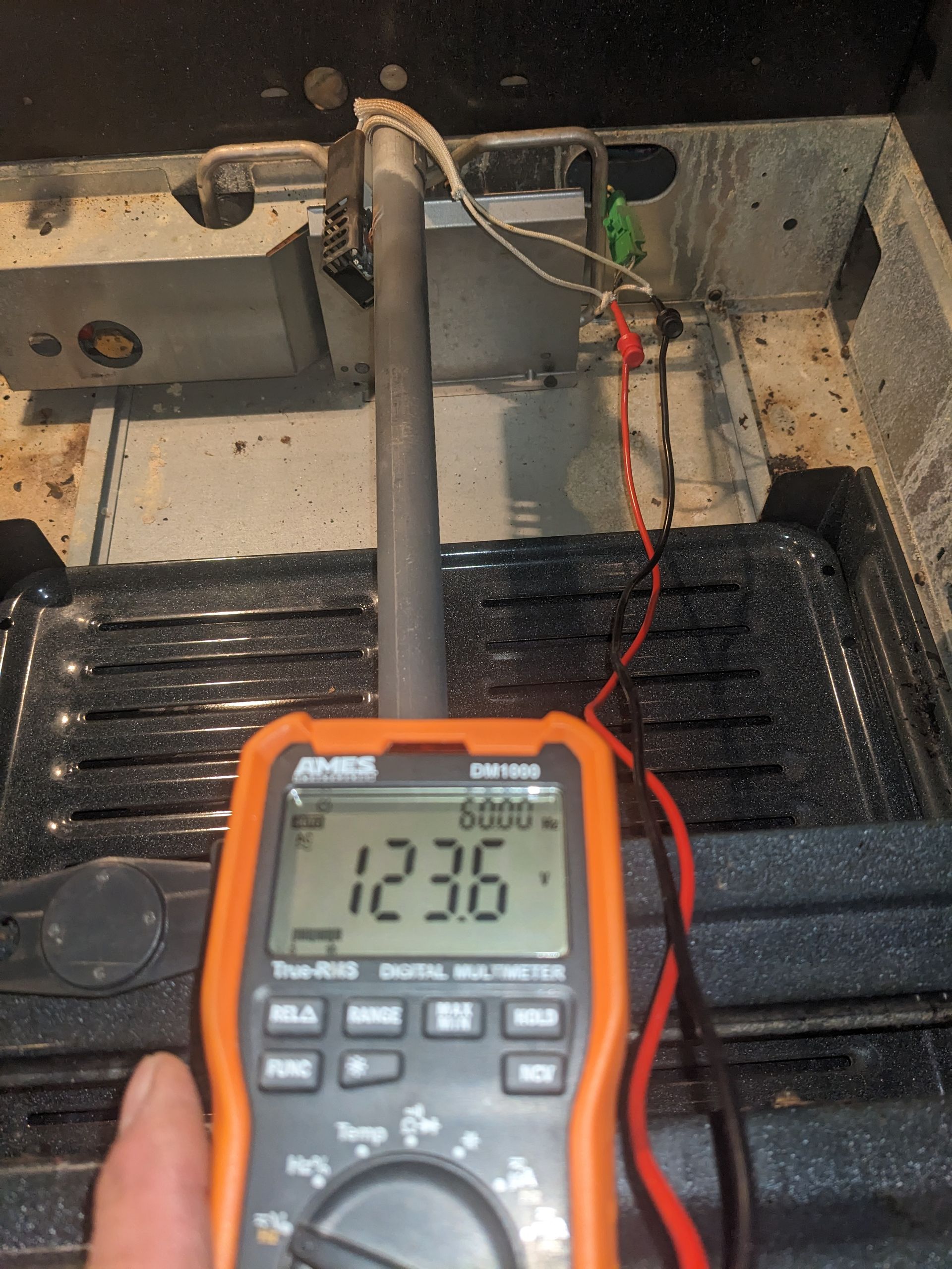 Handheld multimeter measuring 12.36 volts inside an engine compartment with exposed wiring