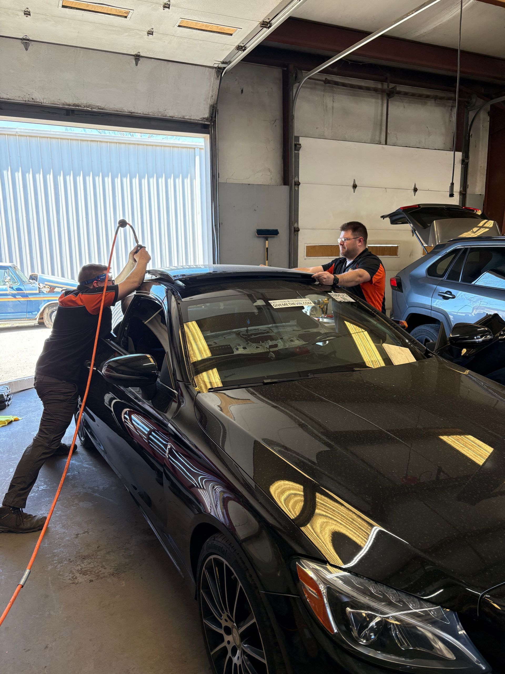 The image depicts two individuals working on a black Mercedes-Benz vehicle, likely performing window tinting or a similar detailing service.