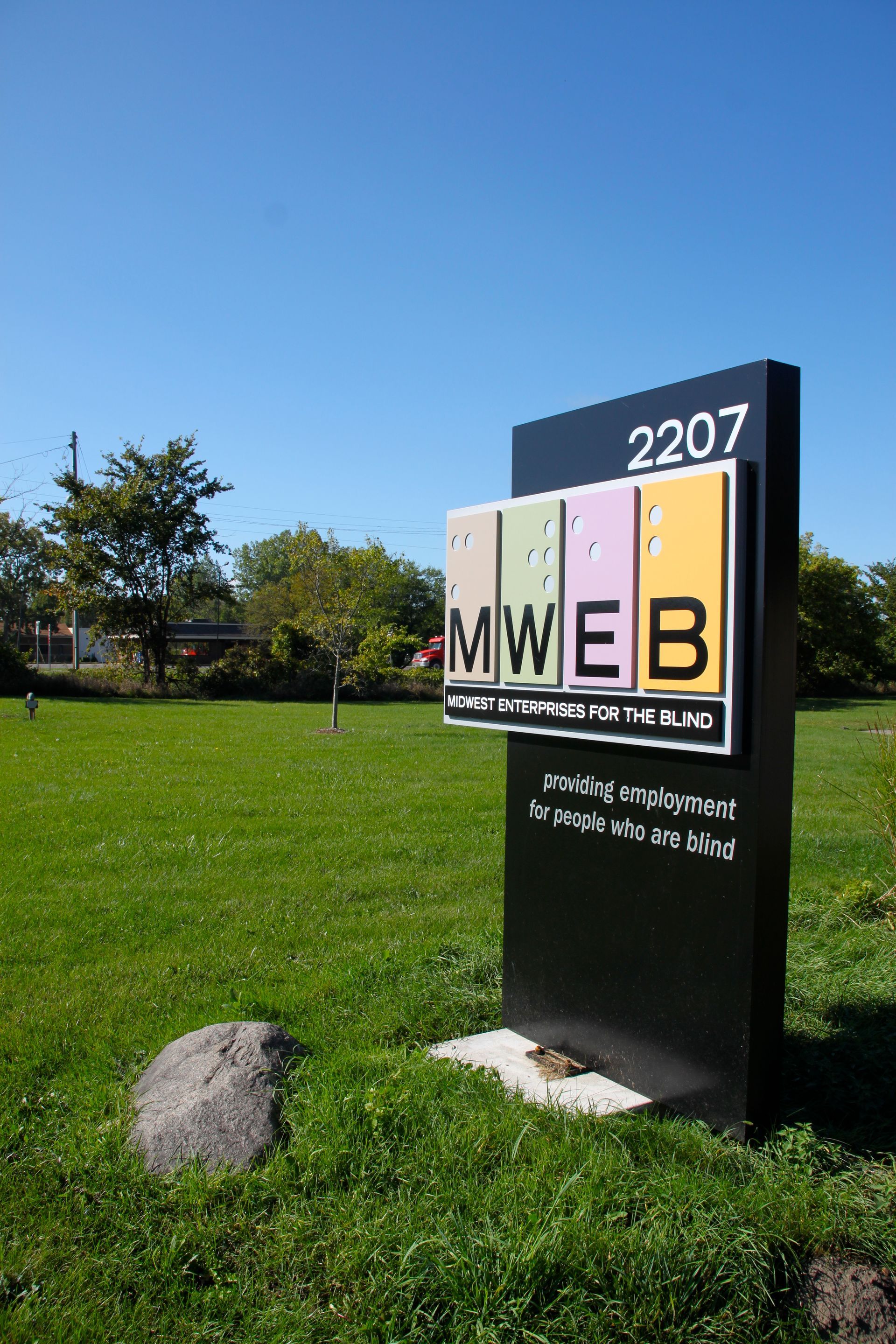 Sign for 2207 MWEB in black with a colorful panel, on green grass under blue sky.