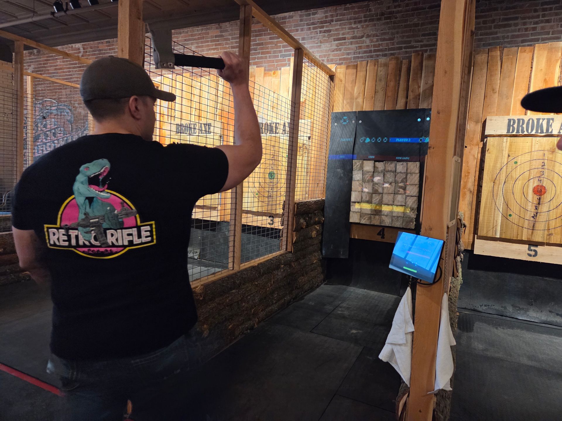 Man aiming an axe in an indoor shooting range lane with wooden partitions and a blue monitor