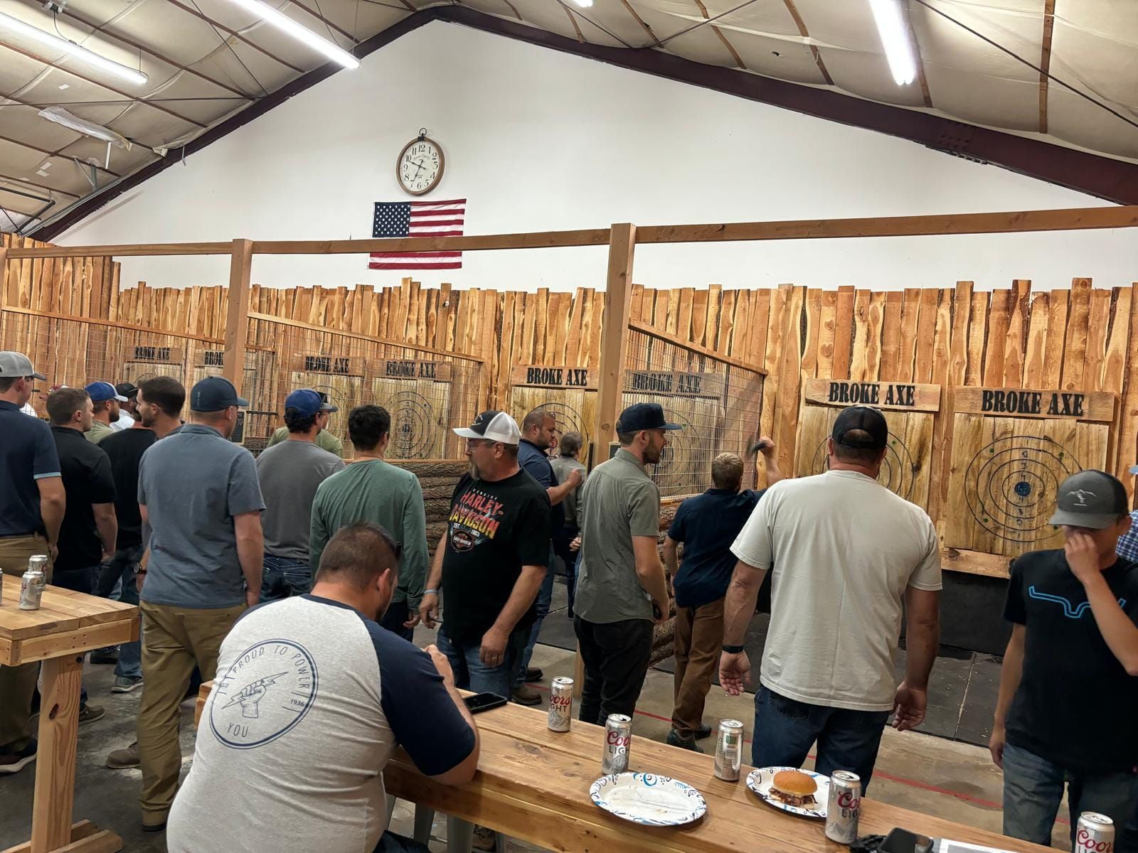 People standing at long tables in a rustic hall, with a US flag and clock on the wall.
