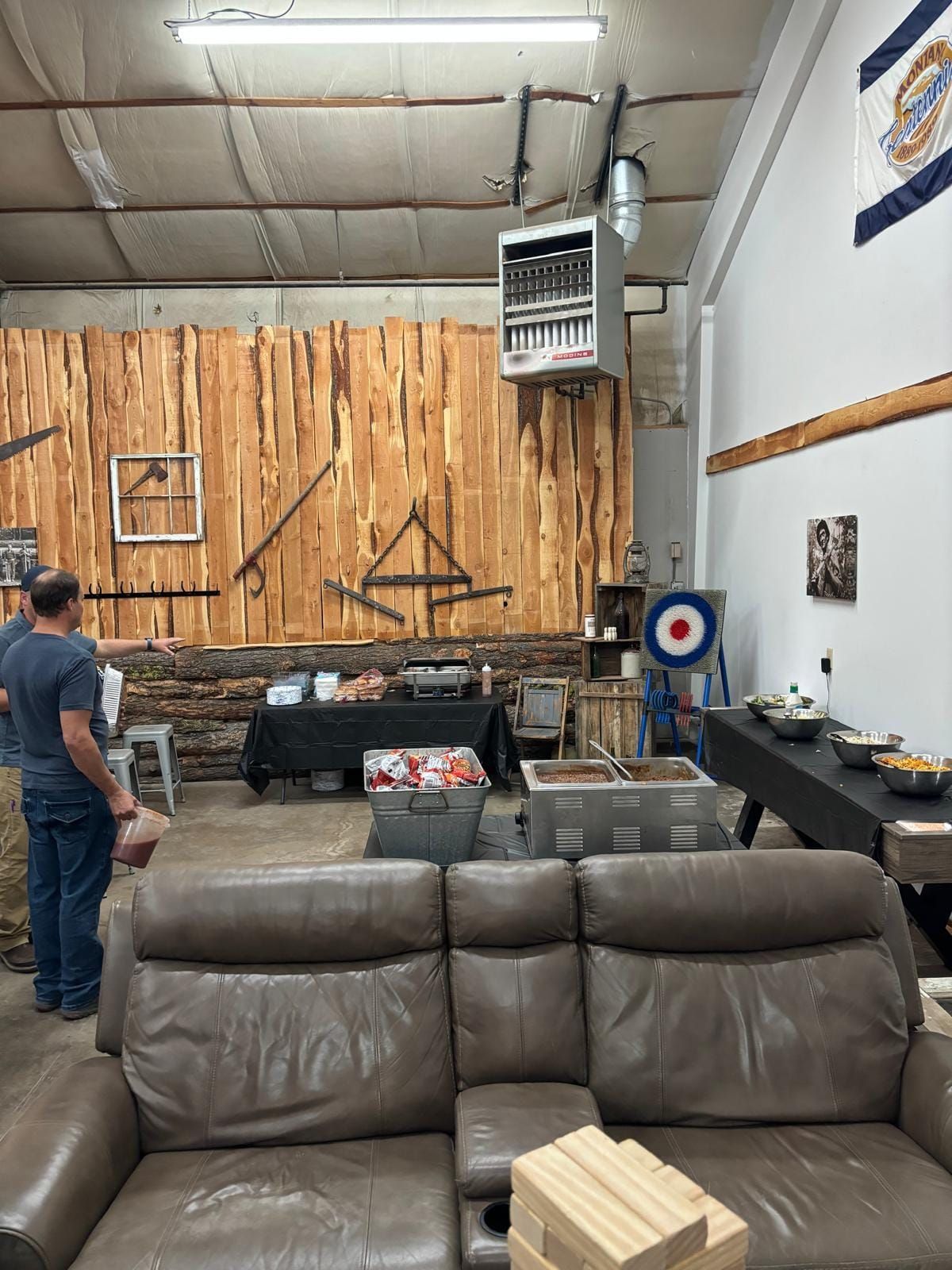 Wood-paneled workshop with hanging tools, a man at left, and a gray couch in the foreground.