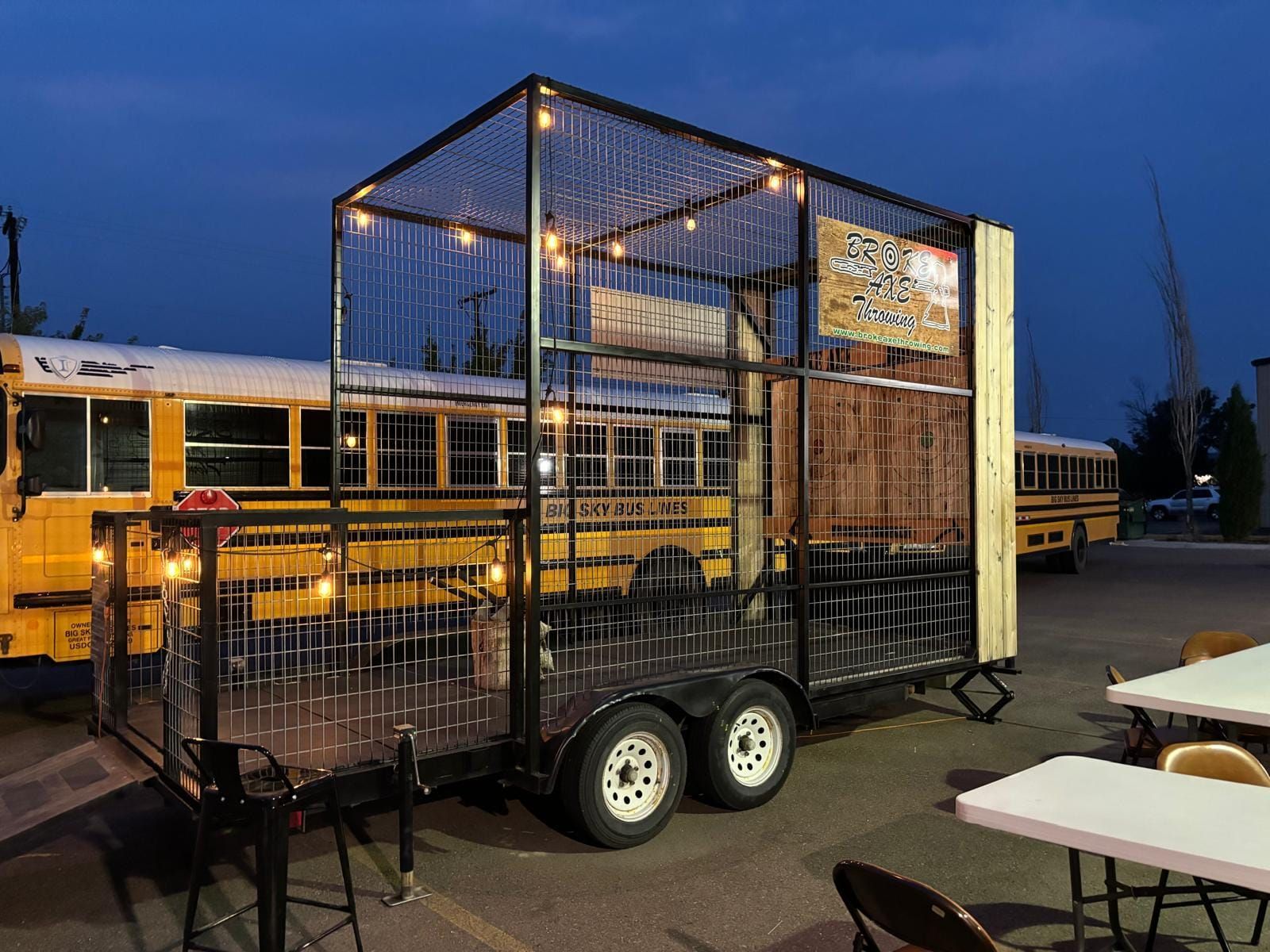 Black metal cage trailer with string lights at dusk, parked beside a yellow school bus.