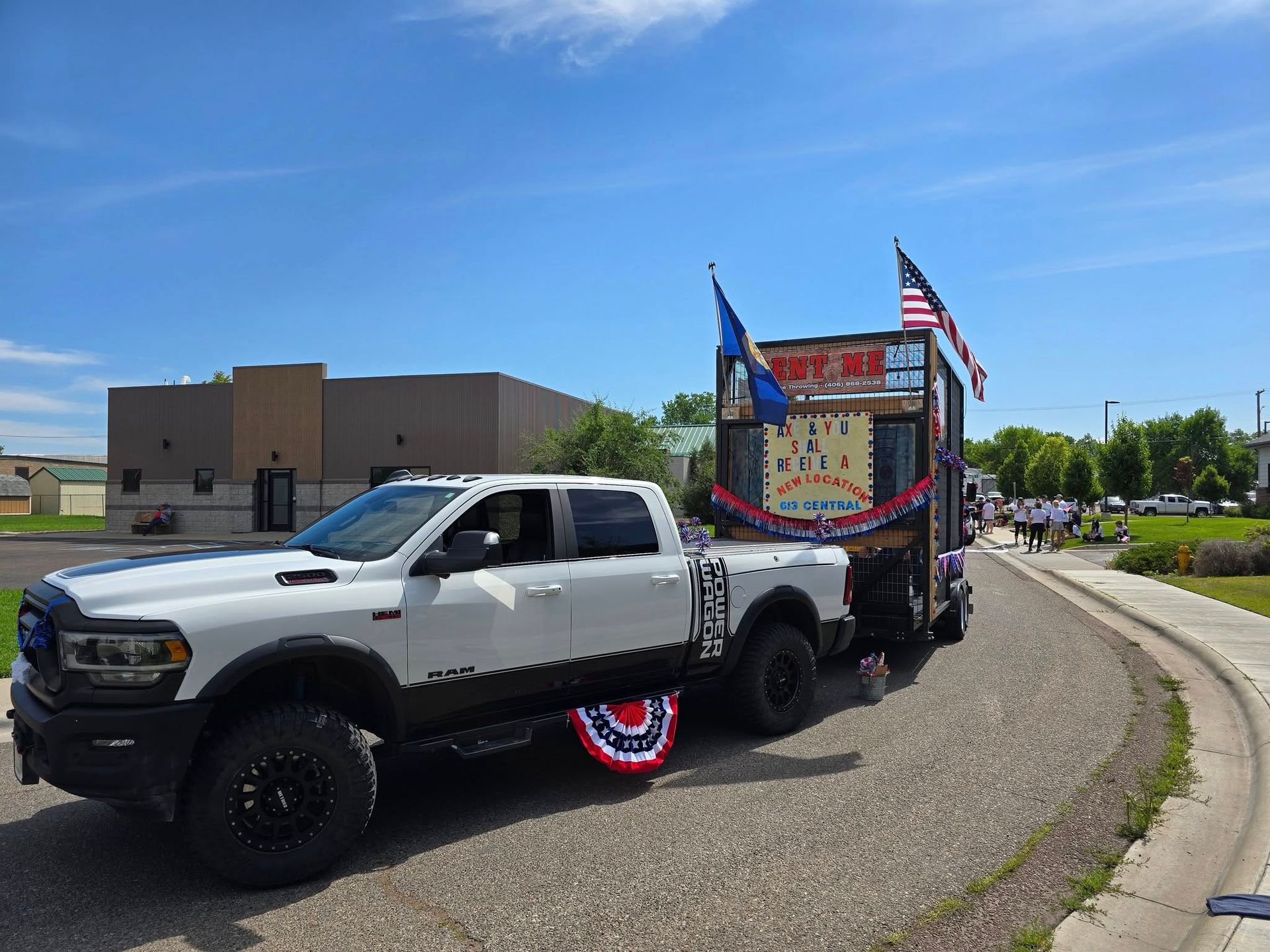 White pickup truck towing a decorated trailer with American flags on a roadside curve under a blue sky