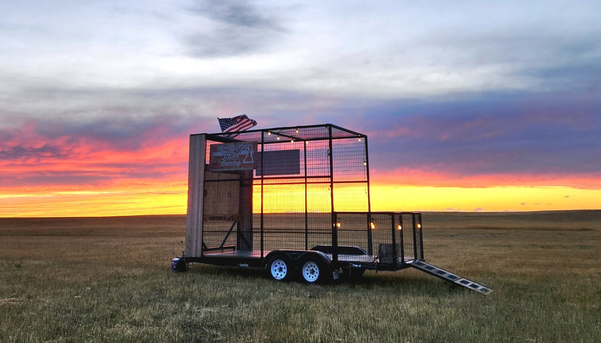 Small trailer-mounted metal cage in a grassy field at sunset, with colorful sky behind it