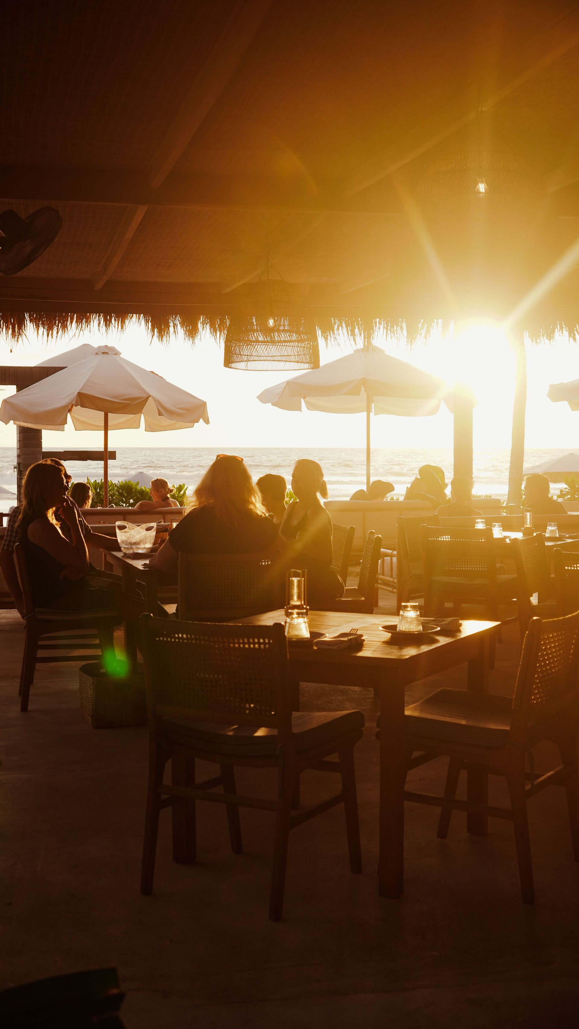 Sunlit beach café with seated patrons, white umbrellas, and tables at sunset