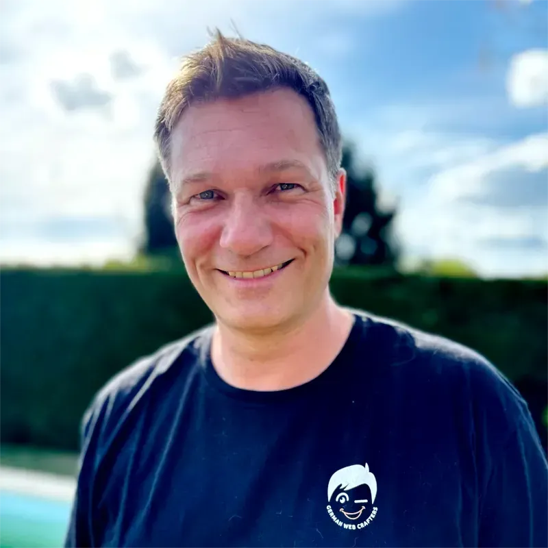 Smiling person in a black T-shirt standing outdoors with a blurred hedge and blue sky background