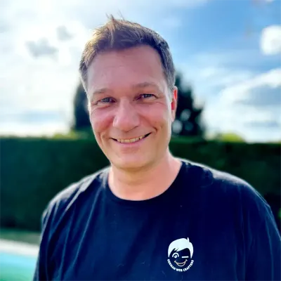 Smiling person in a black T-shirt outdoors, with a blurred hedge and blue sky behind.