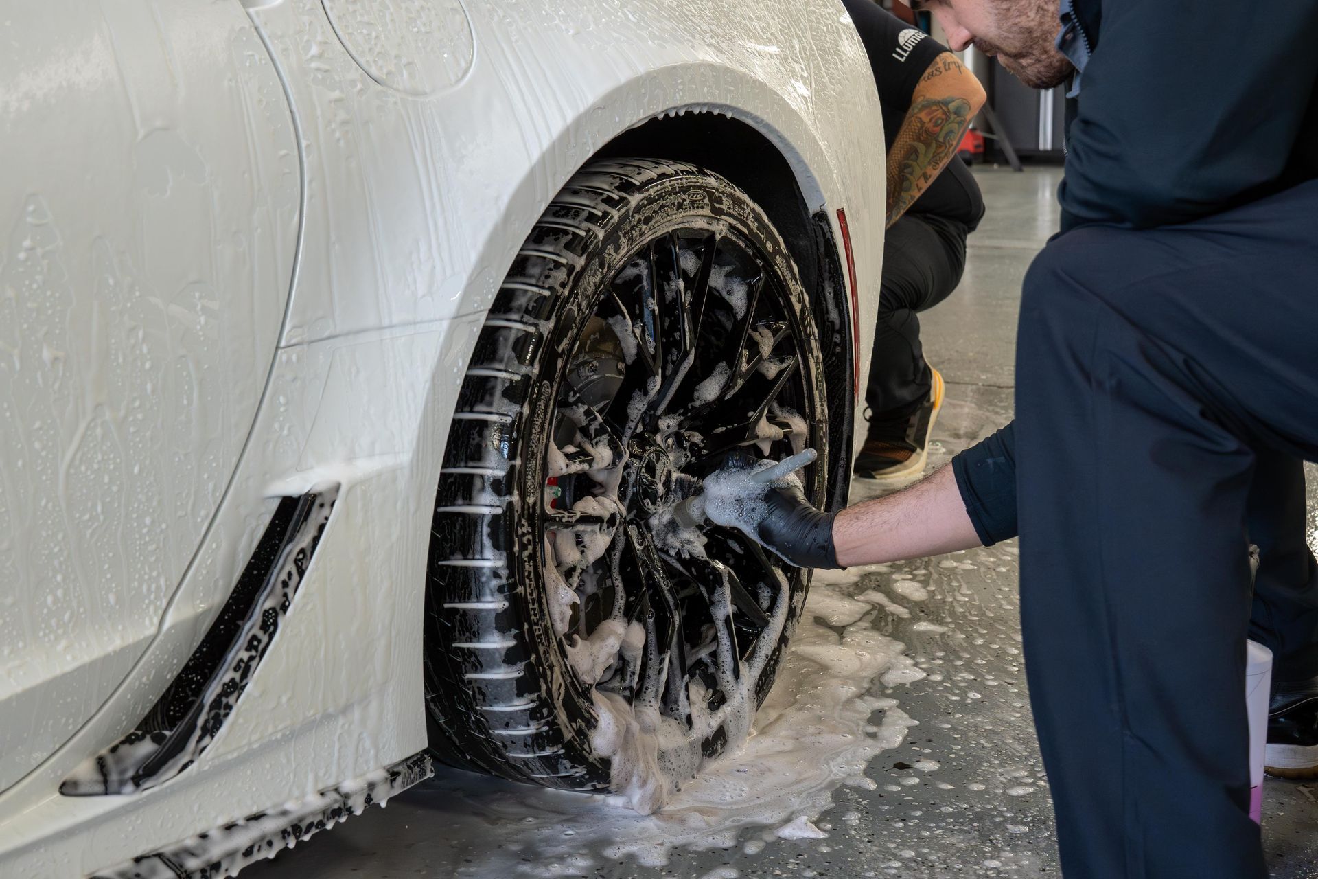 A technician in black uniform cleans a soapy white car wheel with a sponge in an automotive garage.