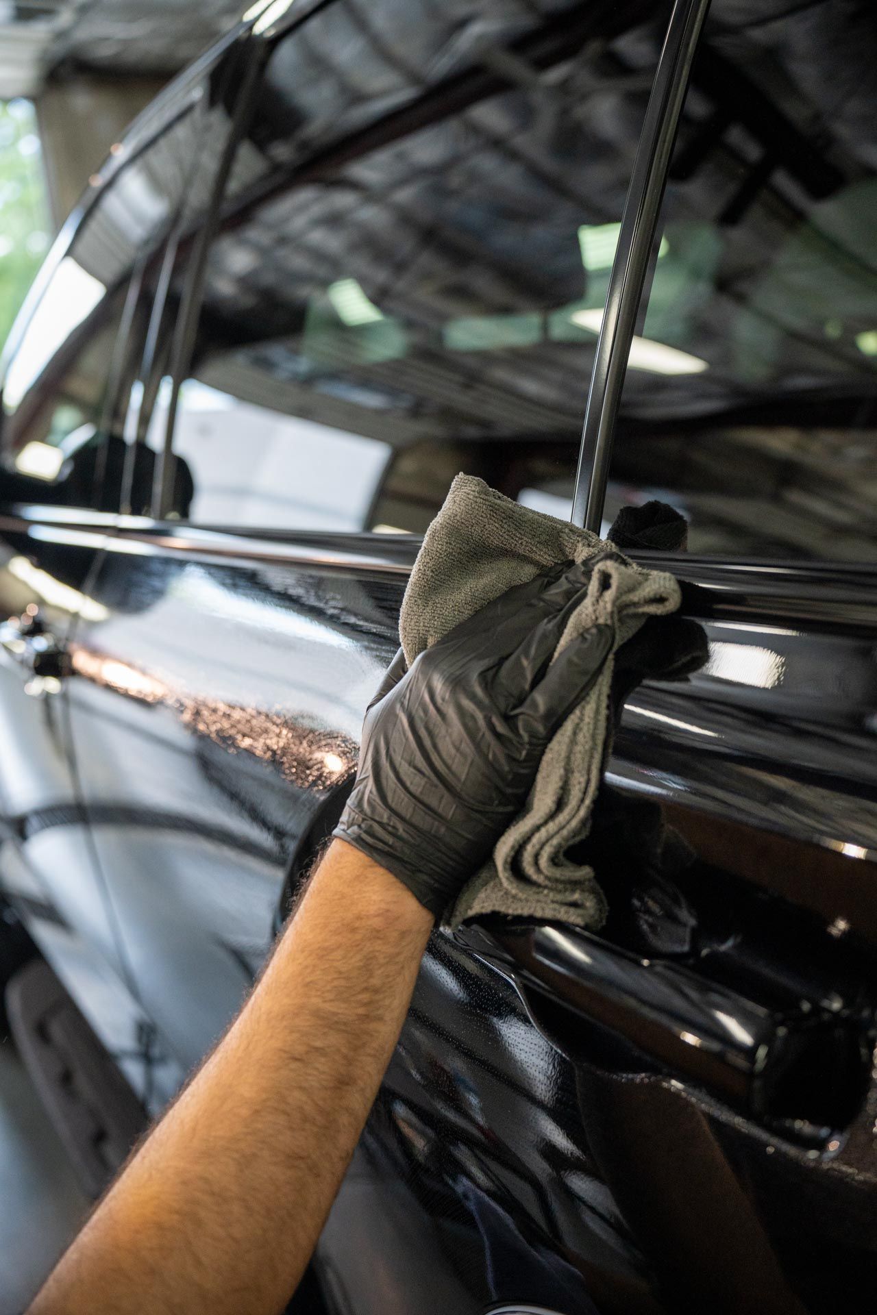A person in a black glove uses a grey cloth to wipe the glossy black exterior of a car door.