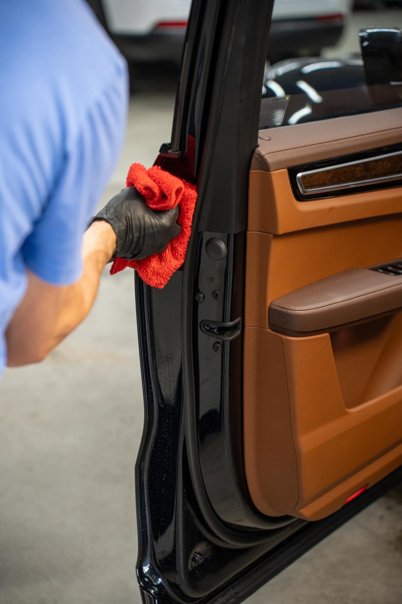 A person wearing a black glove cleans the black door jamb of a vehicle with a red microfiber cloth.