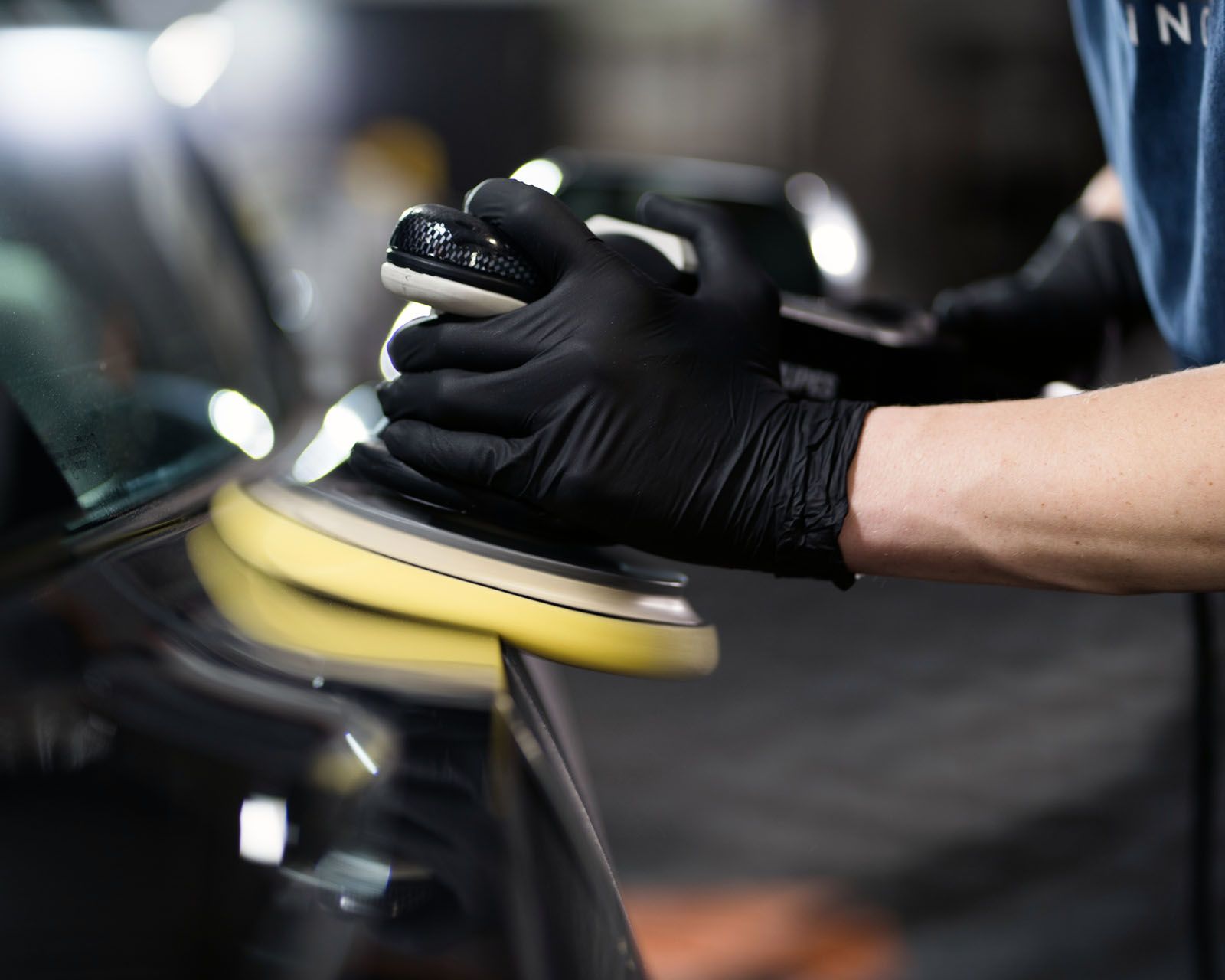 A gloved hand uses a yellow power polisher to buff a black car's exterior in a garage setting.