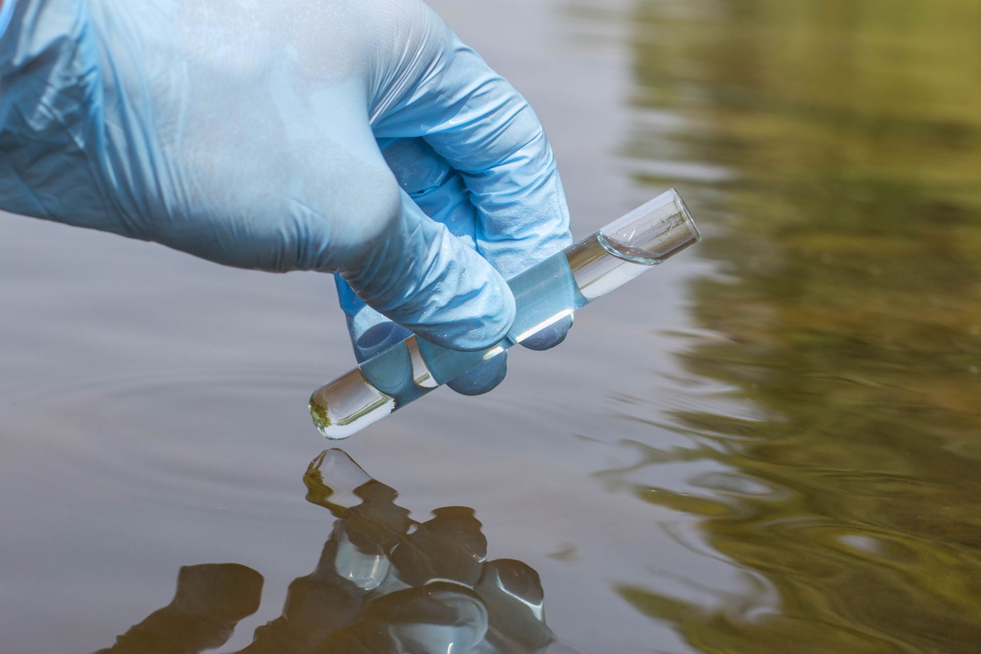Gloved hand collecting a water sample in a vial from a body of water outdoors. Gloved hand collecting a water sample in a vial from a body of water outdoors.