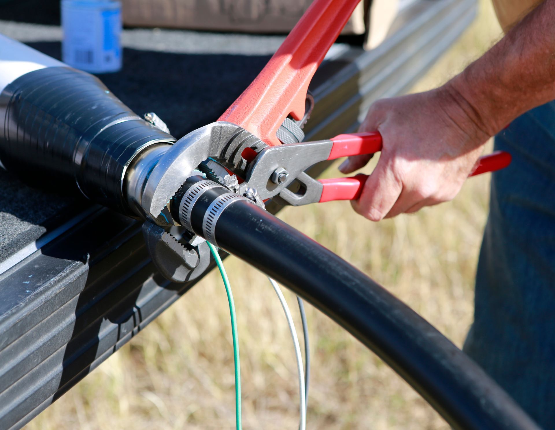 Person uses pliers to tighten a clamp on a black hose attached to a white pipe outdoors.